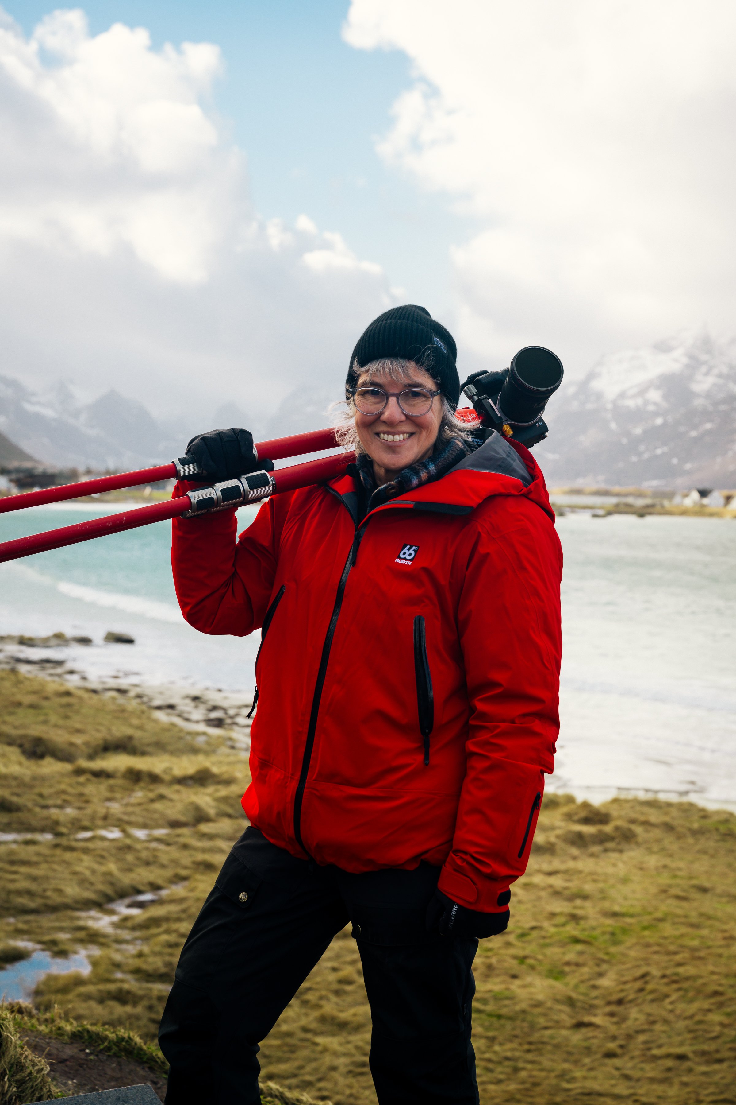 Smiling woman with curly gray hair and glasses, wearing an orange rain jacket and gloves, holding a camera outdoors in front of a waterfall.