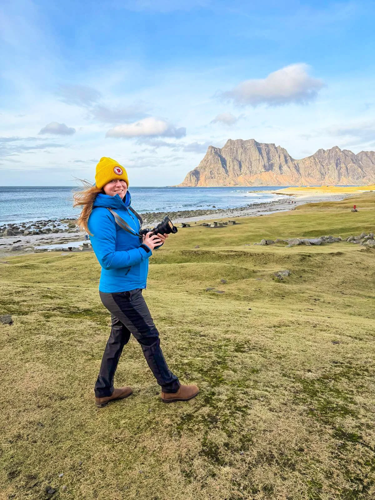A smiling woman with red hair holding a Nikon camera in a bright room, with artwork and equipment in the background.
