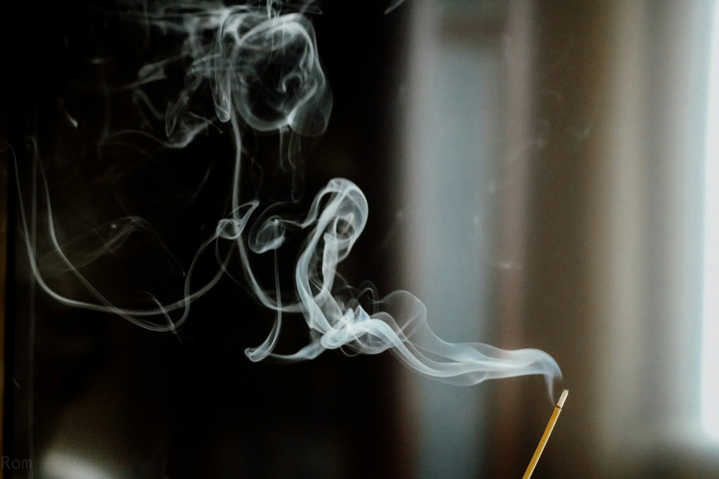White smoke rising from a lit incense stick against a dark background, with some light coming from the right side.