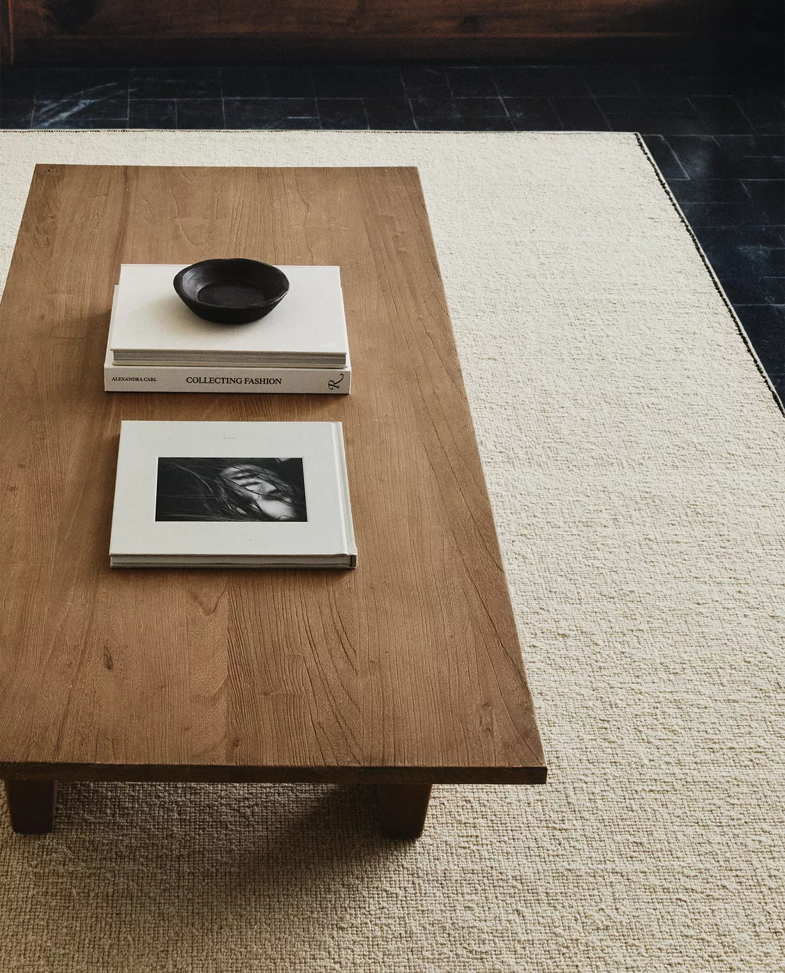 A wooden coffee table on a beige rug with two books and a small black dish on top.