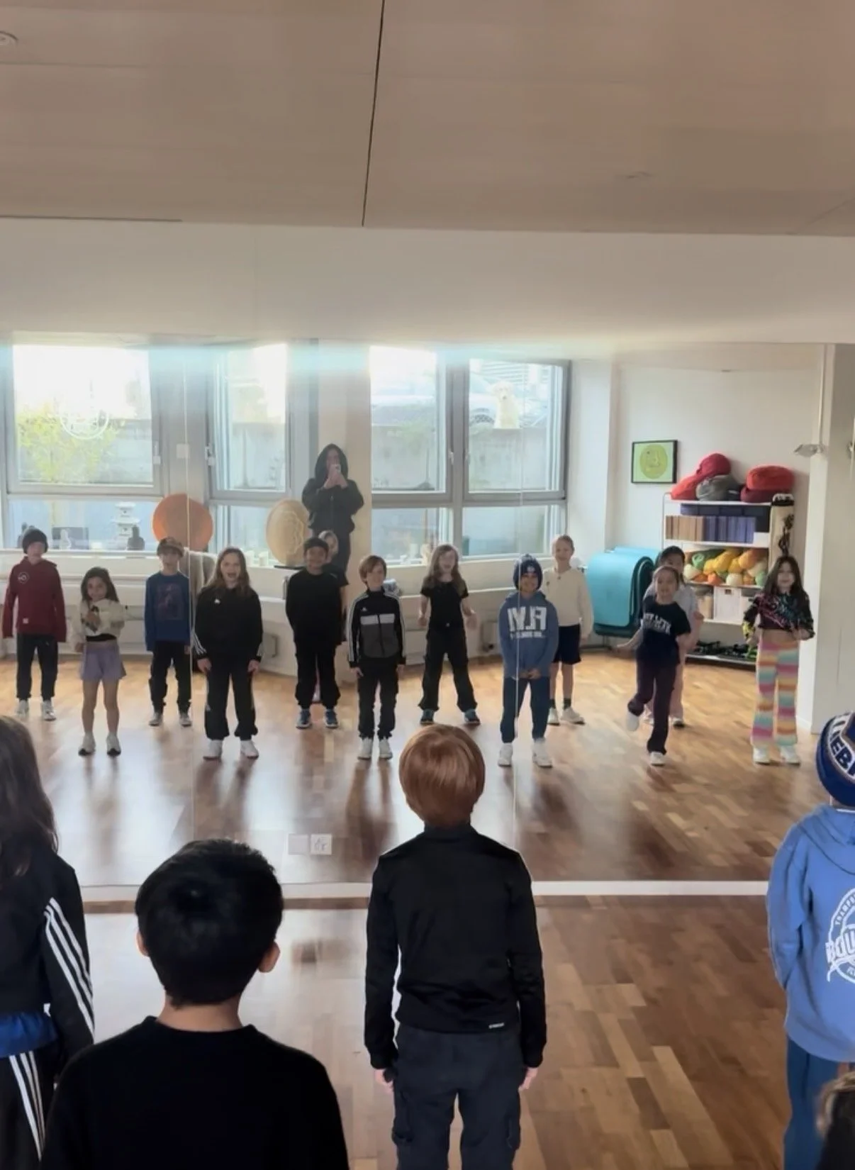 Eine Gruppe von Kindern steht im Raum, einige schauen und einige tanzen oder bewegen sich, während eine erwachsene Person im Hintergrund steht und Fotos macht.