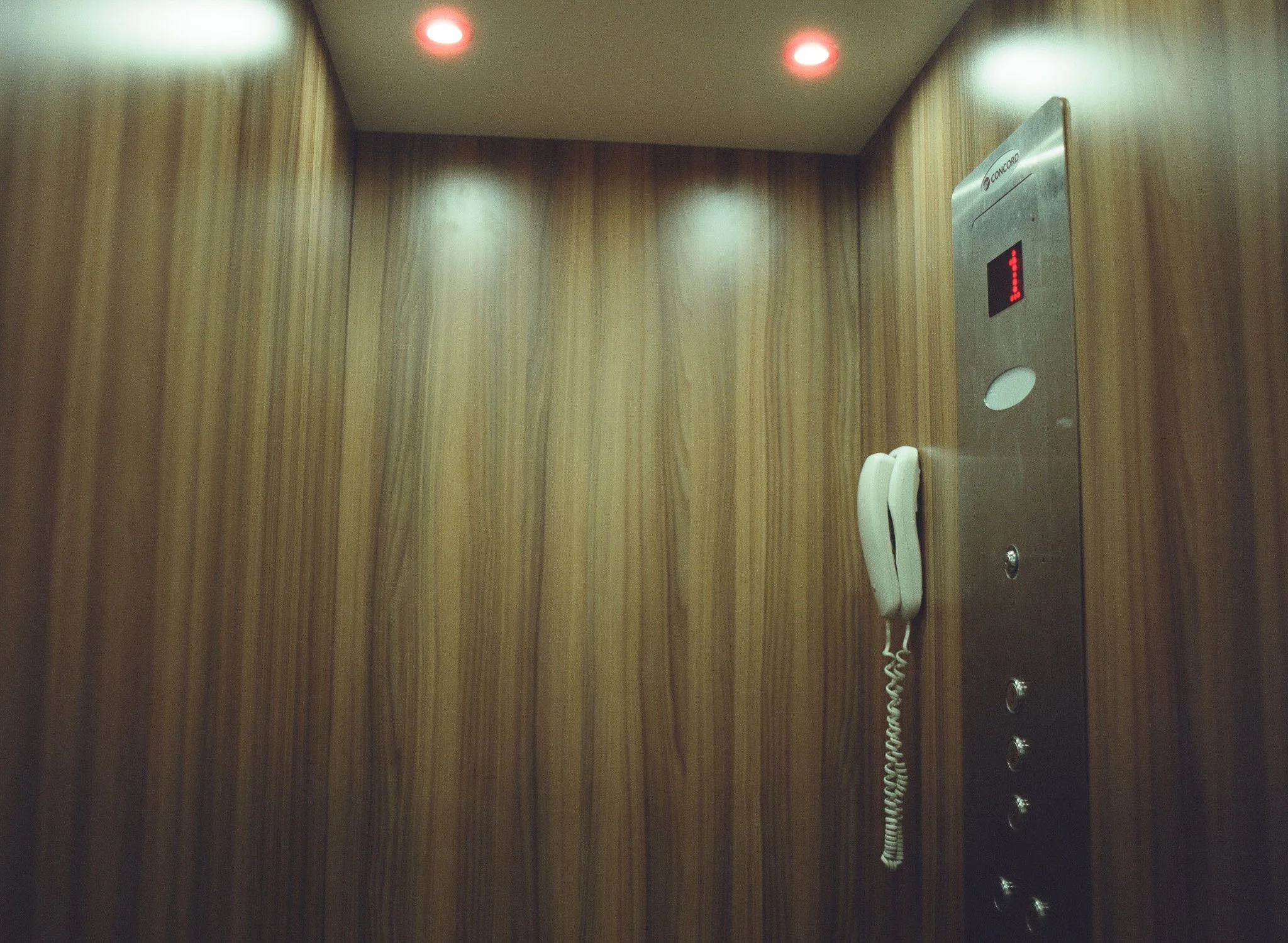 Interior of an elevator with wooden paneling, red ceiling lights, a white telephone mounted on the wall, and a control panel with an illuminated number 1.