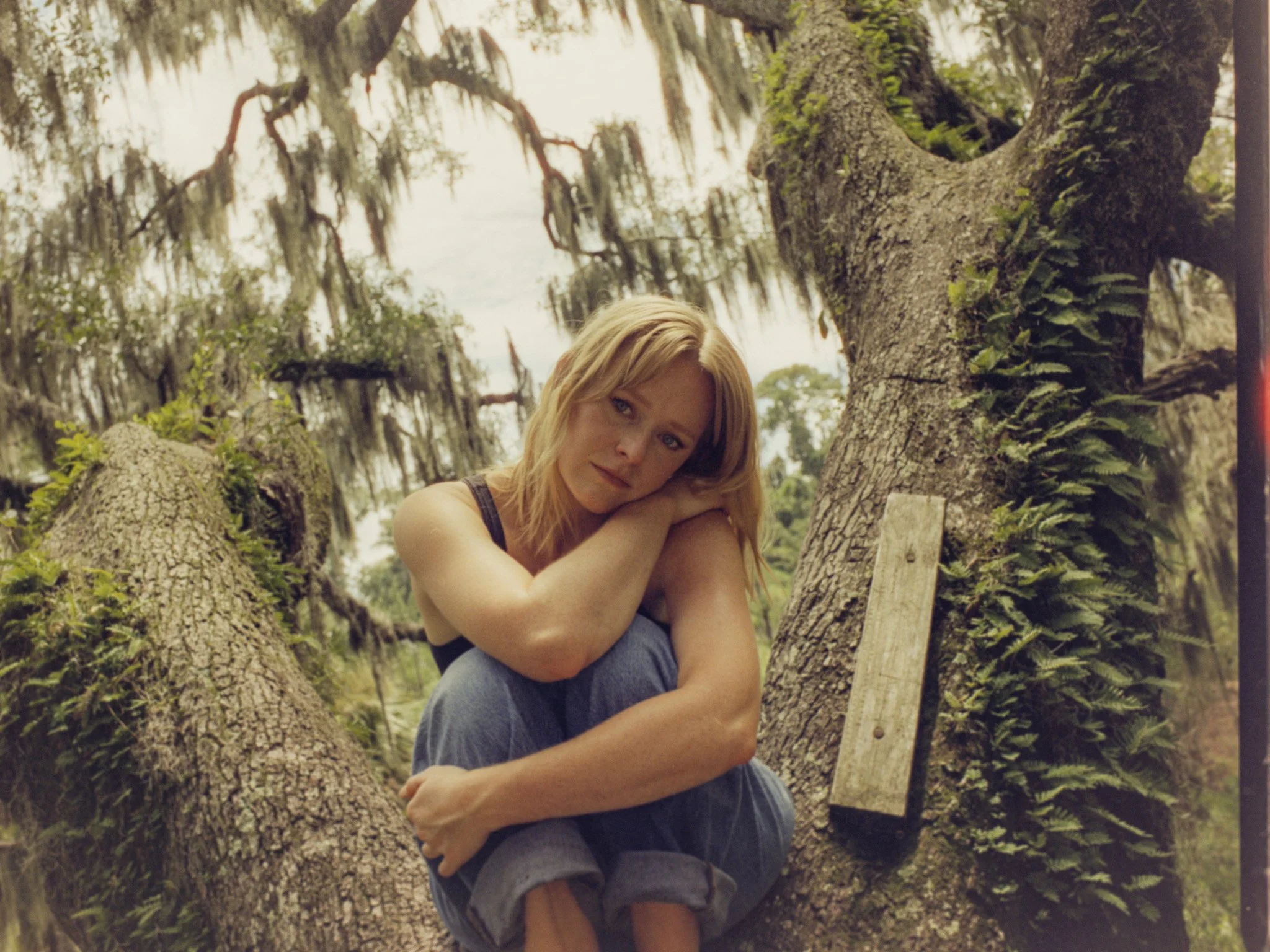 A woman with blonde hair sitting on a tree branch, resting her head on her arms, surrounded by moss-covered tree branches and foliage.