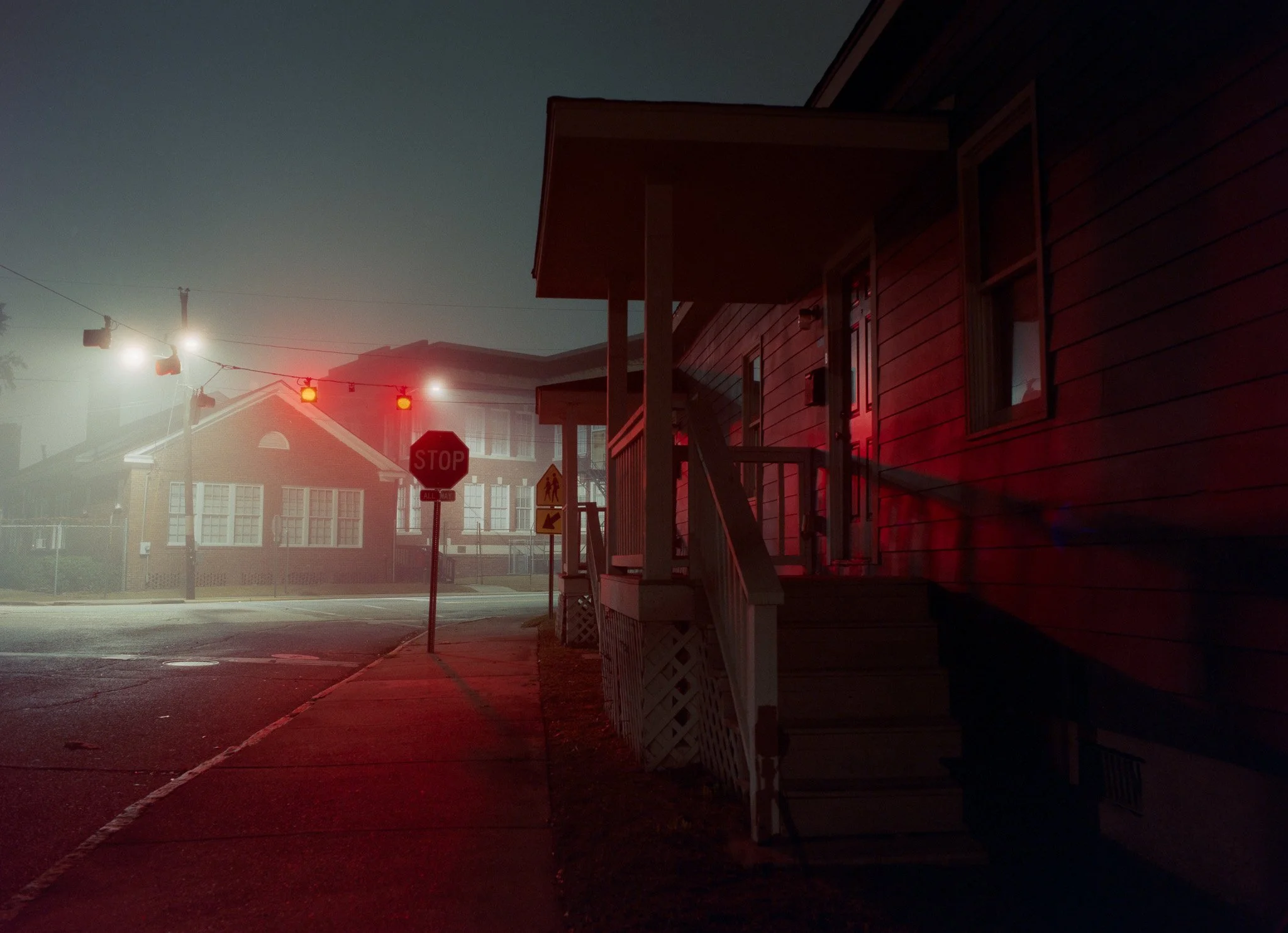 A residential street corner at night with traffic lights showing red, a stop sign, and a building with a wooden deck in the foreground. The scene is dimly lit with some street light and building windows.