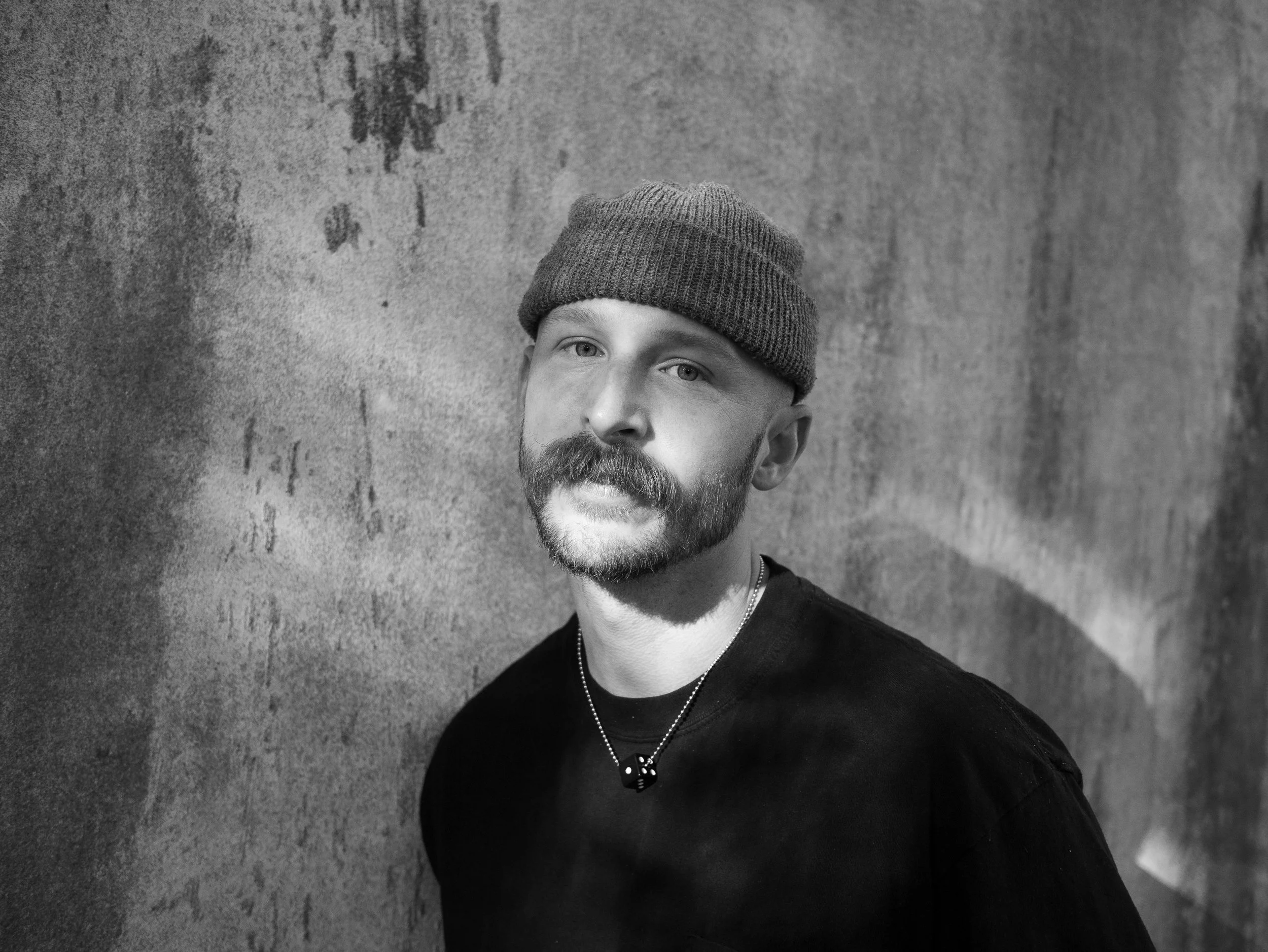 Black and white portrait of a man with a beard and mustache, wearing a beanie and a necklace, posing against a textured wall.