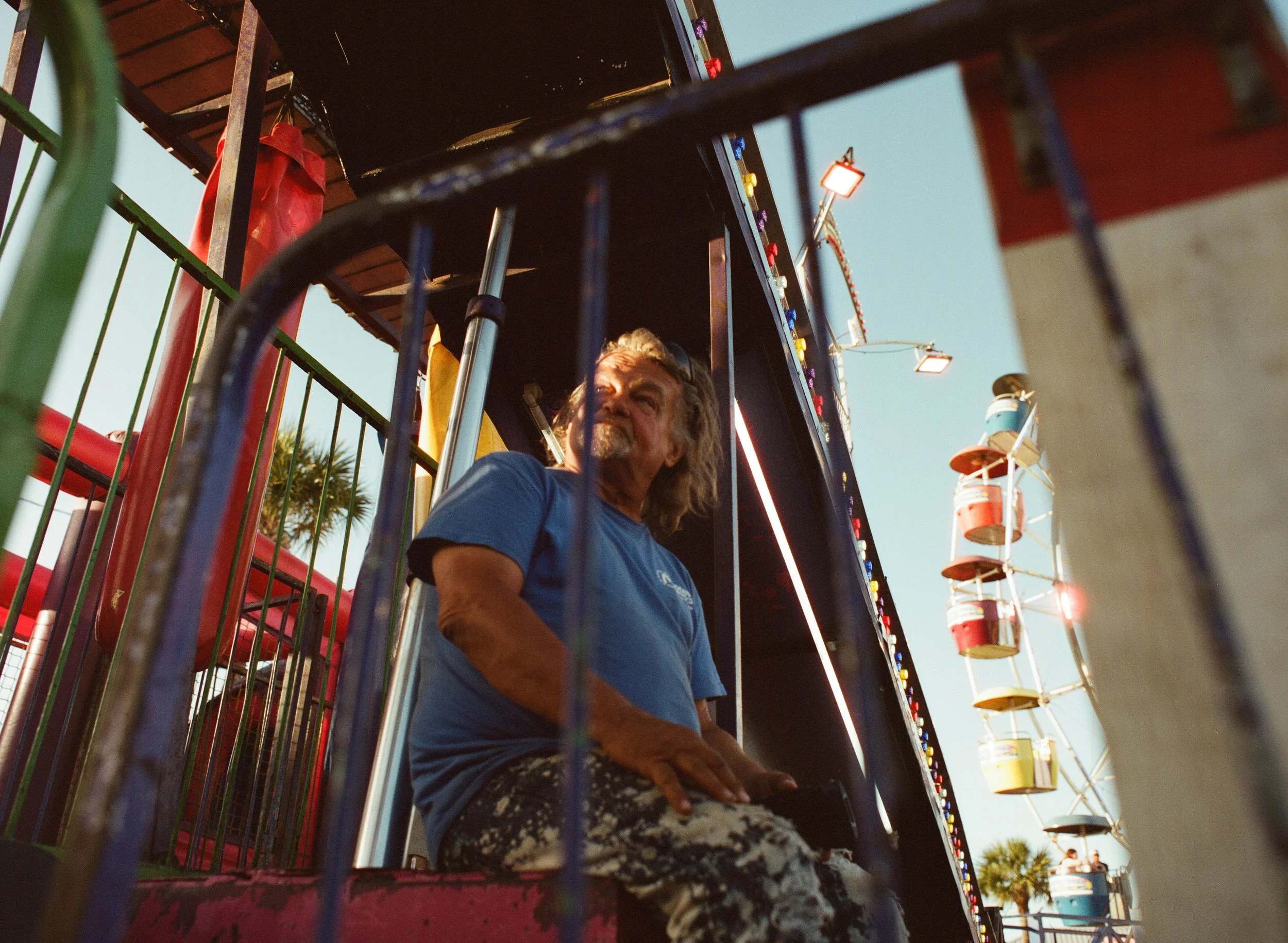 A man with long hair and a beard sitting on a carnival ride, seen through colorful metal bars, with a Ferris wheel in the background and palm trees, under a clear blue sky.