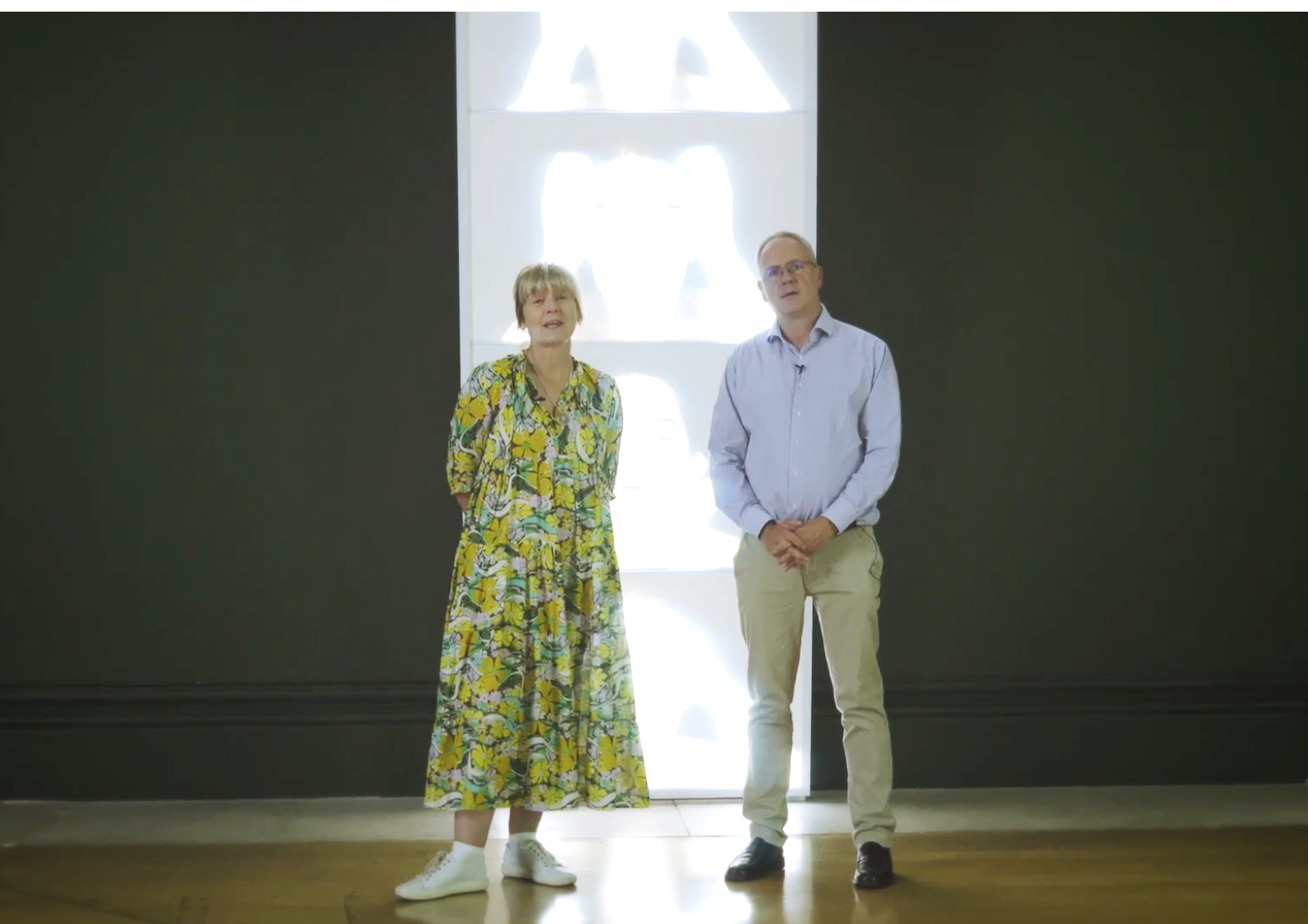 A woman in a colorful floral dress and a man in a light blue shirt and beige pants standing indoors in front of a dark wall with a bright, abstract artwork behind them.