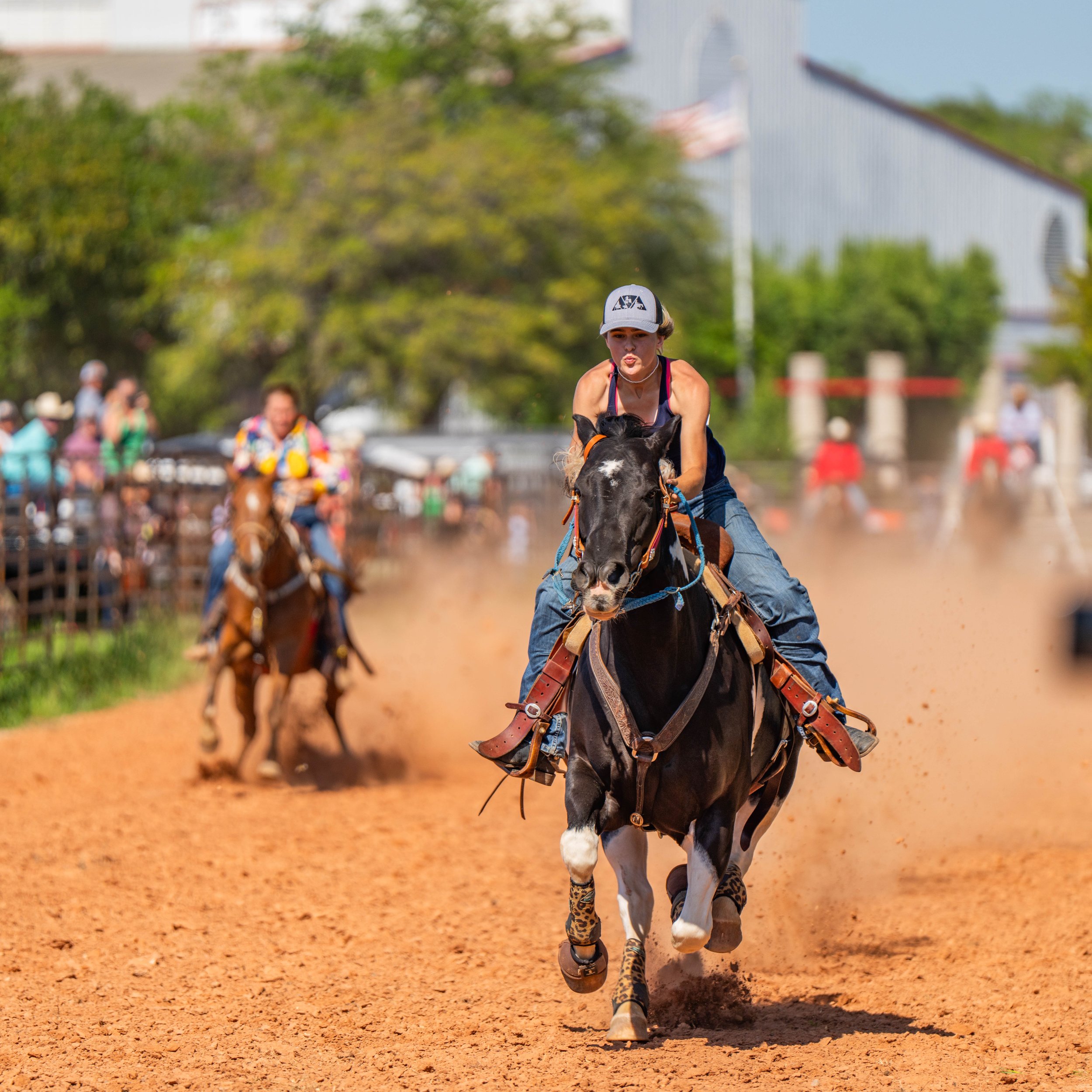 A woman riding a black and white horse at a rodeo, with other riders and a crowd in the background on a dusty dirt track.