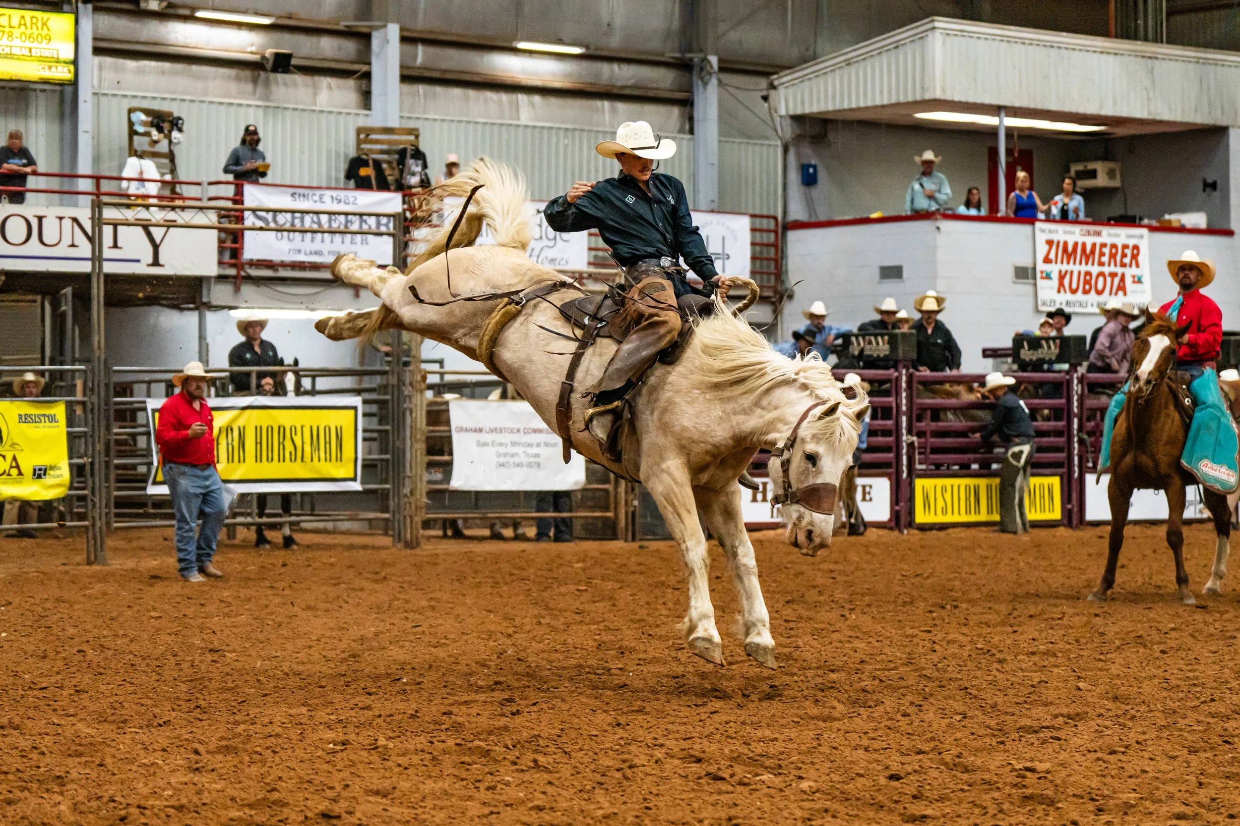 A cowboy riding a bucking horse inside an arena at a rodeo, with spectators and announcers in the background.