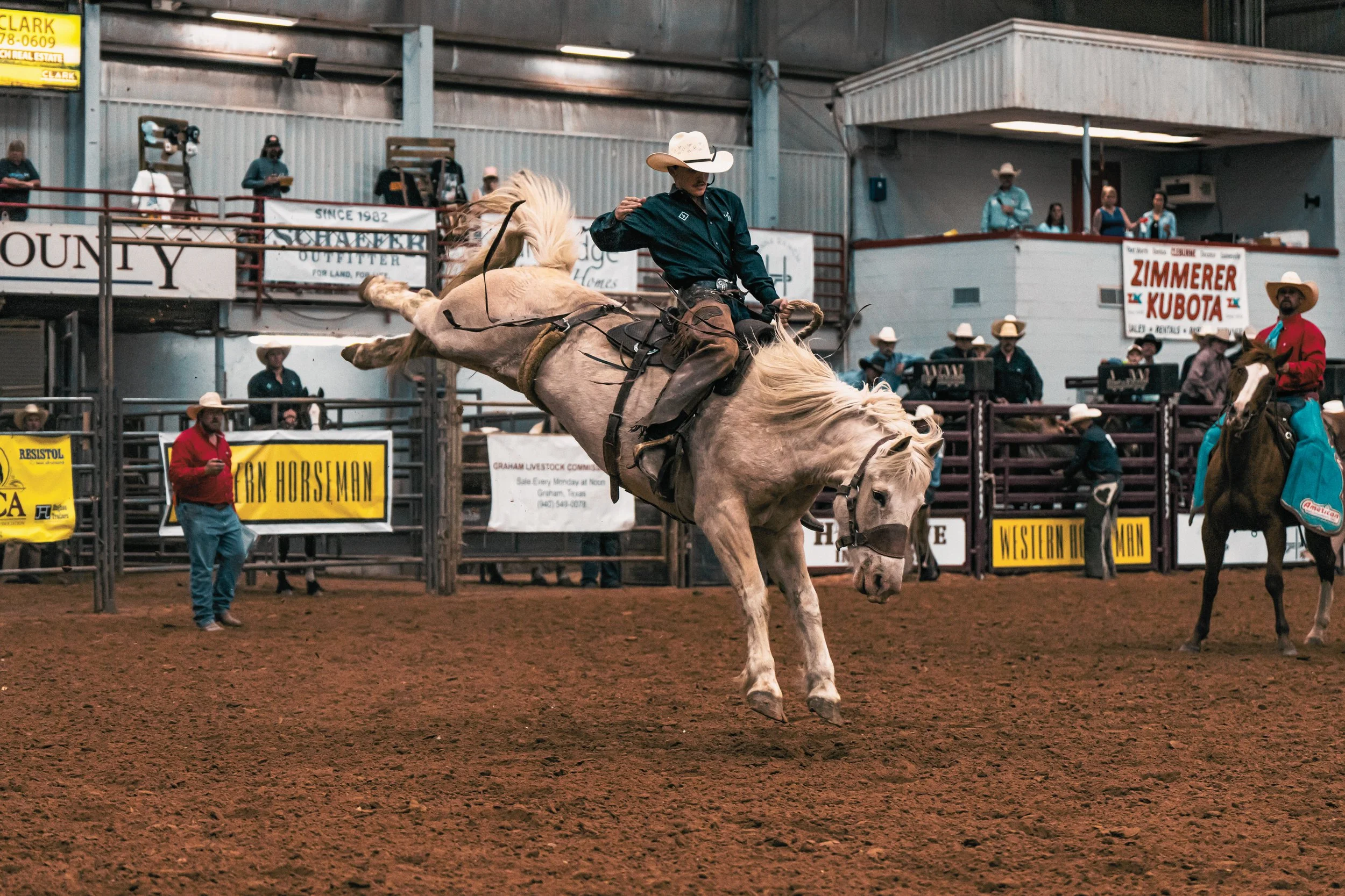 A cowboy riding a bucking horse inside an arena at a rodeo, with spectators and announcers in the background.
