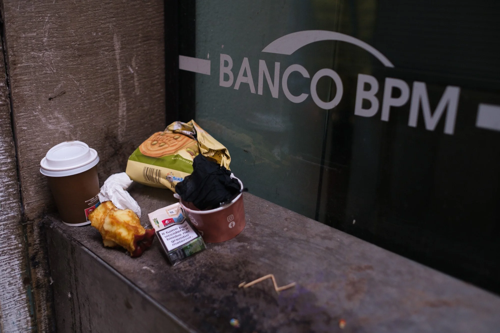 Resti di una colazione abbandonati su un davanzale — caffè, cornetto, sigarette — davanti a una vetrina a Venezia