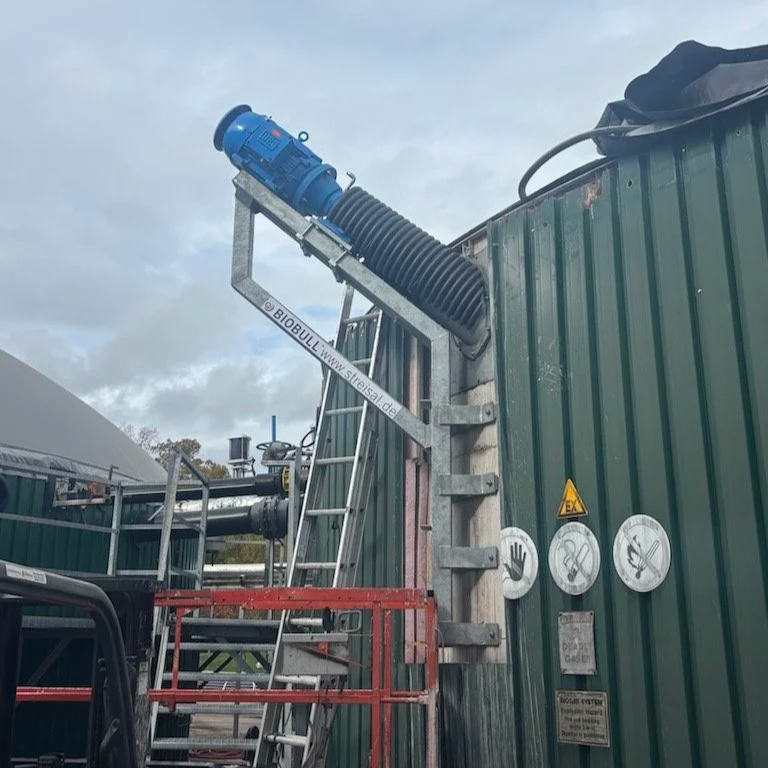 Agitator motor and mounting frame on the outside of an anaerobic digester tank with green siding removed where install is taking place, scaffolding and ladders in place.