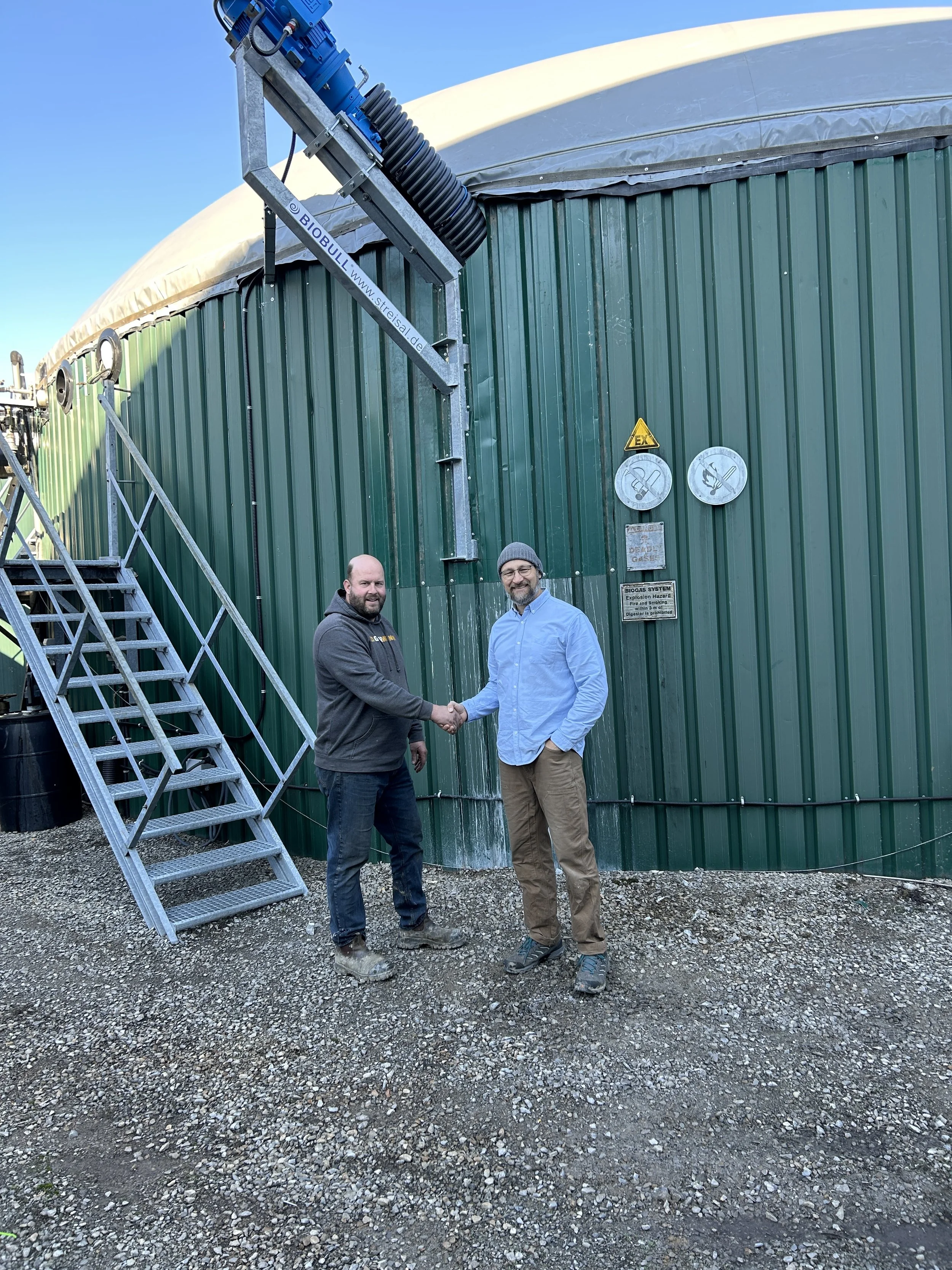 Tom Ferencevic, Fitec CEO, and Bayview Flowers Biogas Operator shaking hands in front of green digester tank. Newly installed agitator above them.