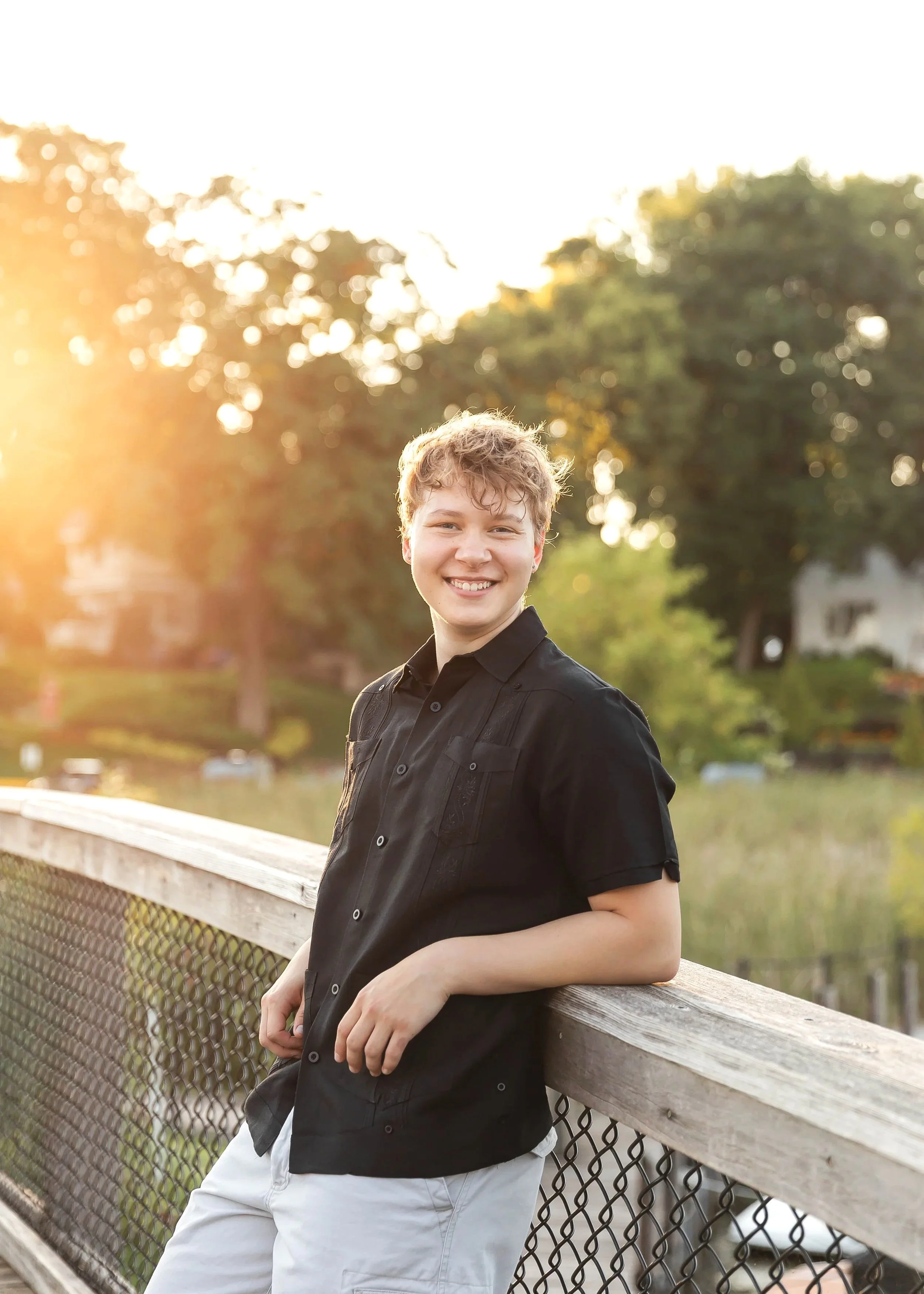 A young man with curly blond hair smiling, leaning on a wooden railing in an outdoor park or nature setting during sunset, with trees and houses in the background.