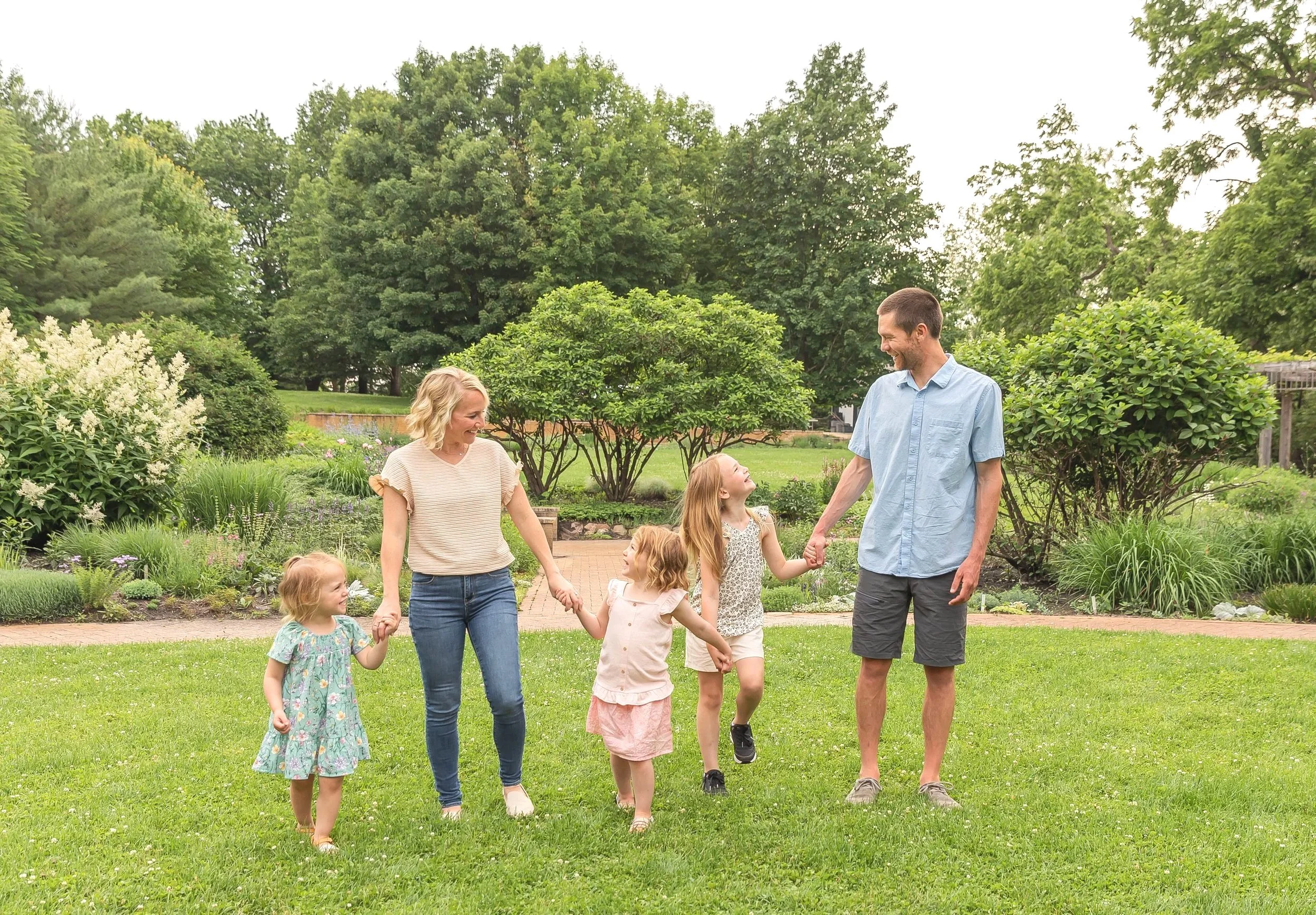 Family of six walking and holding hands in a park with lush green trees and flowering plants.