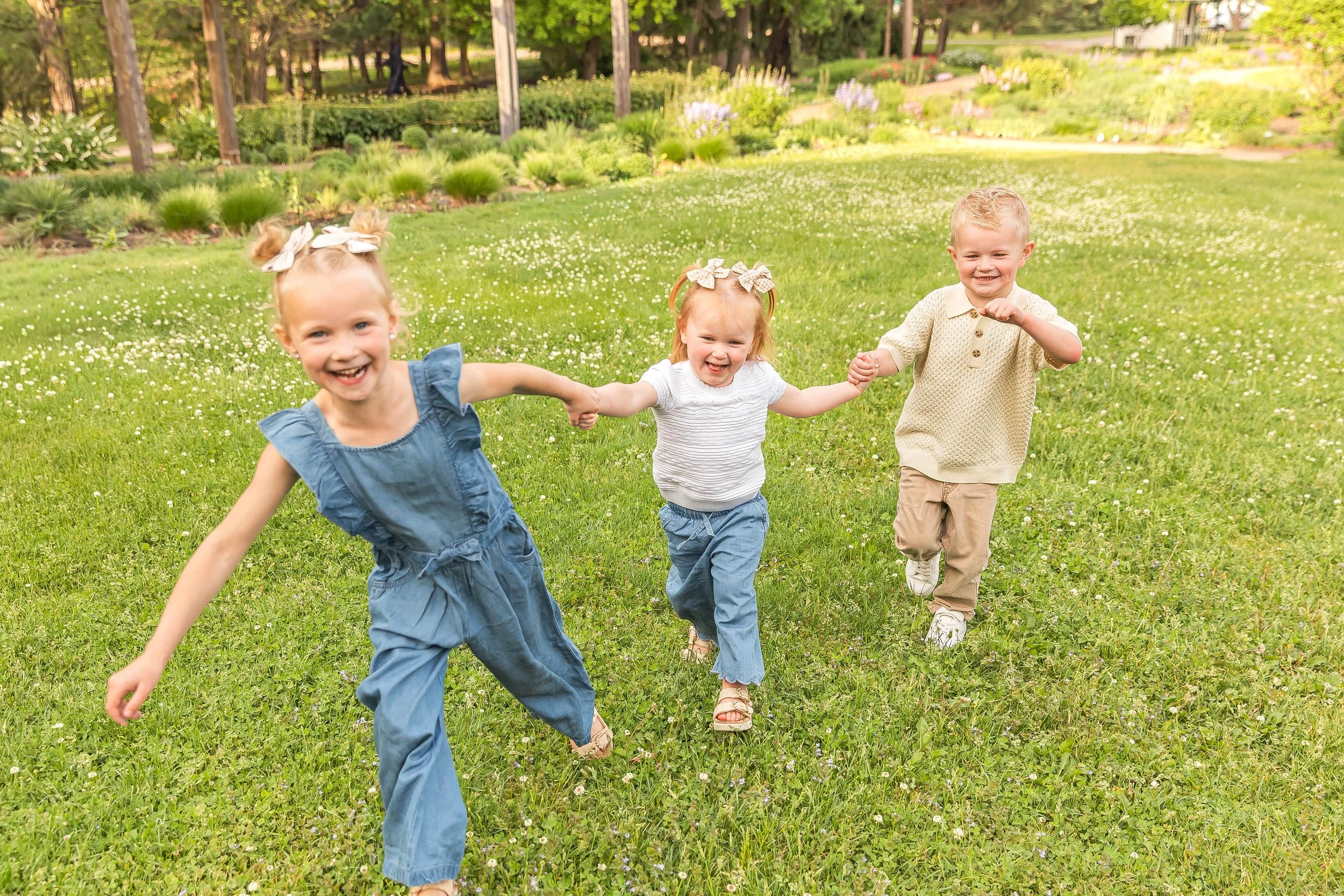 Three young children holding hands and happily running through a grassy field. Family photos at Noerenberg Gardens in Orono, MN.