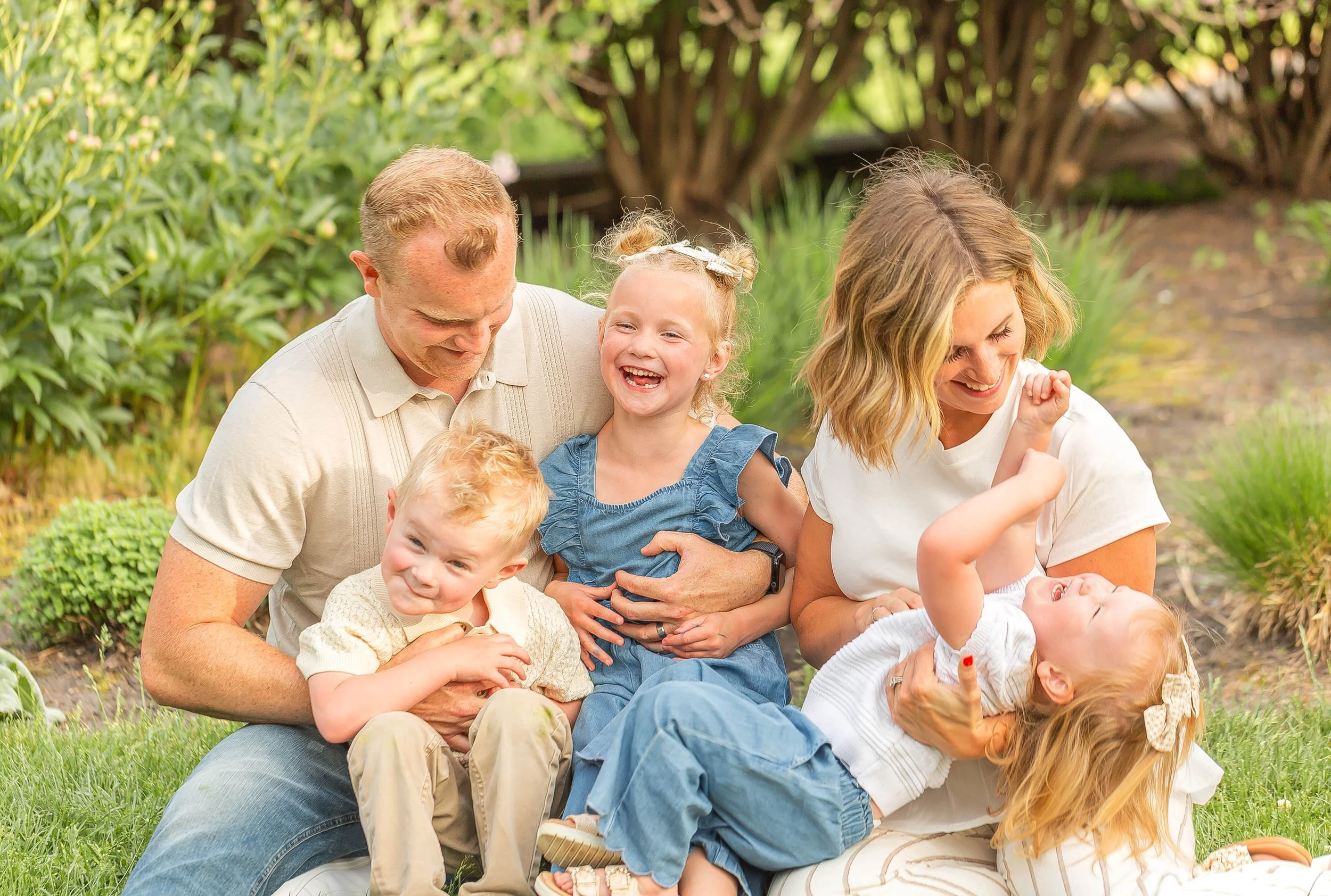Family sitting on grass in a garden, smiling and laughing together, with trees and plants in the background. Family photos at Noerenberg Gardens in Orono, MN.