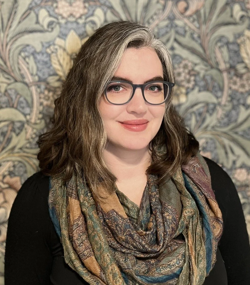 A woman with light skin and shoulder-length wavy brown hair, wearing glasses, a patterned scarf, and a black top, smiling in front of a floral patterned wallpaper background.