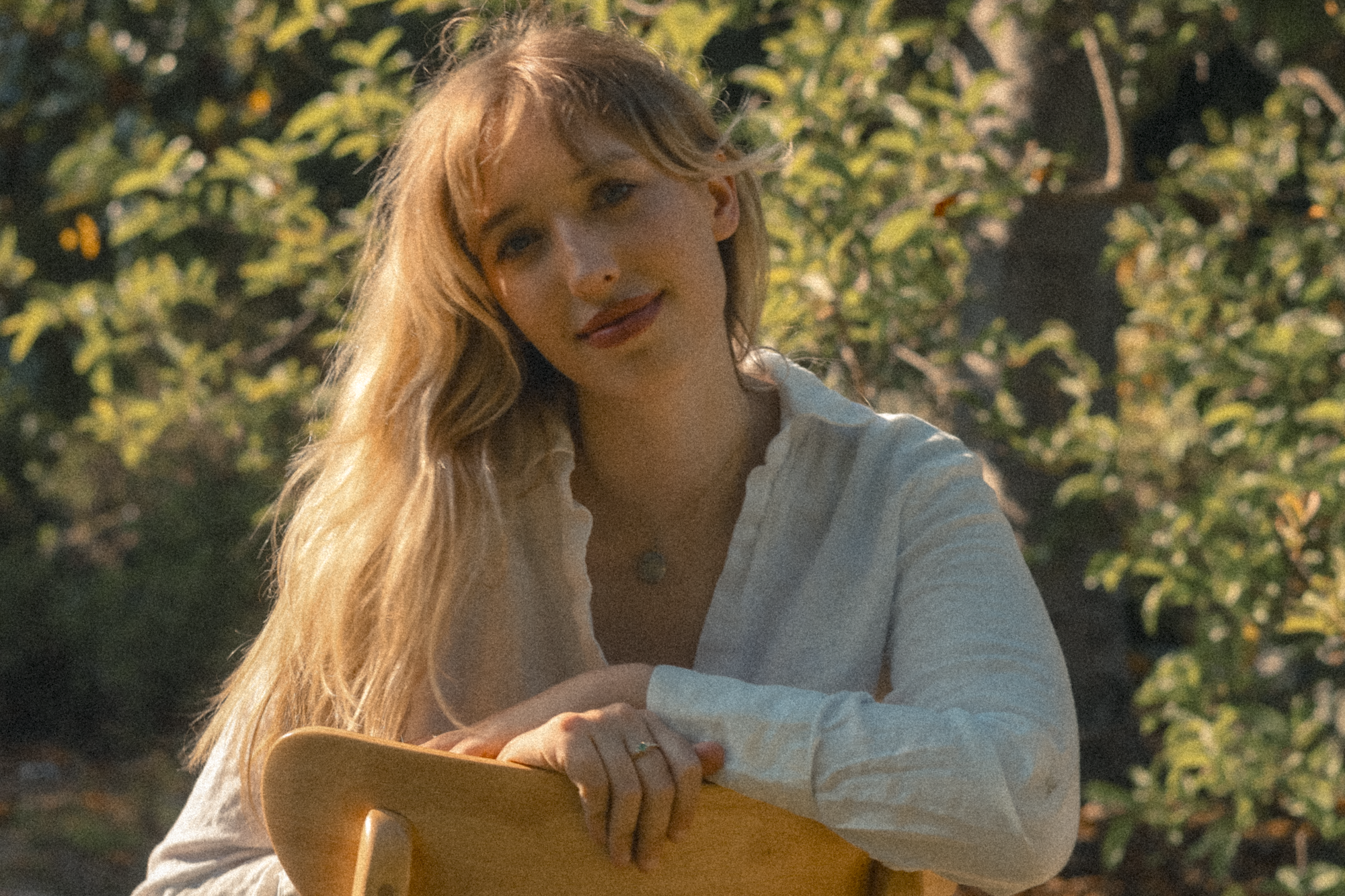 A young woman with long blonde hair, wearing a white shirt, stands outdoors during daytime, with greenery and sunlight in the background.
