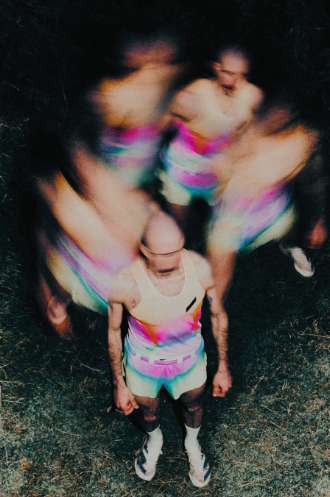 Multiple blurred images of a woman with a shaved head and glasses, wearing a colorful striped dress, standing on a dark surface.