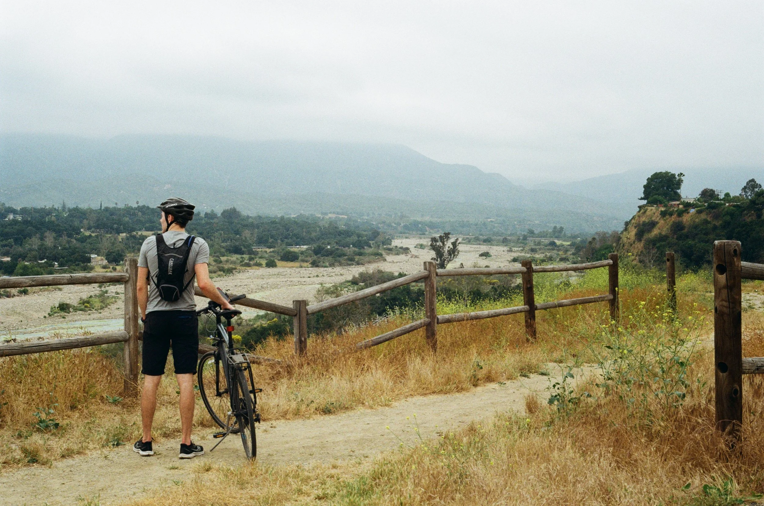 A person wearing a helmet, grey t-shirt, black shorts, and sneakers standing on a trail, holding a mountain bike with a scenic landscape of mountains, trees, and a cloudy sky in the background, with a wooden fence in the foreground.