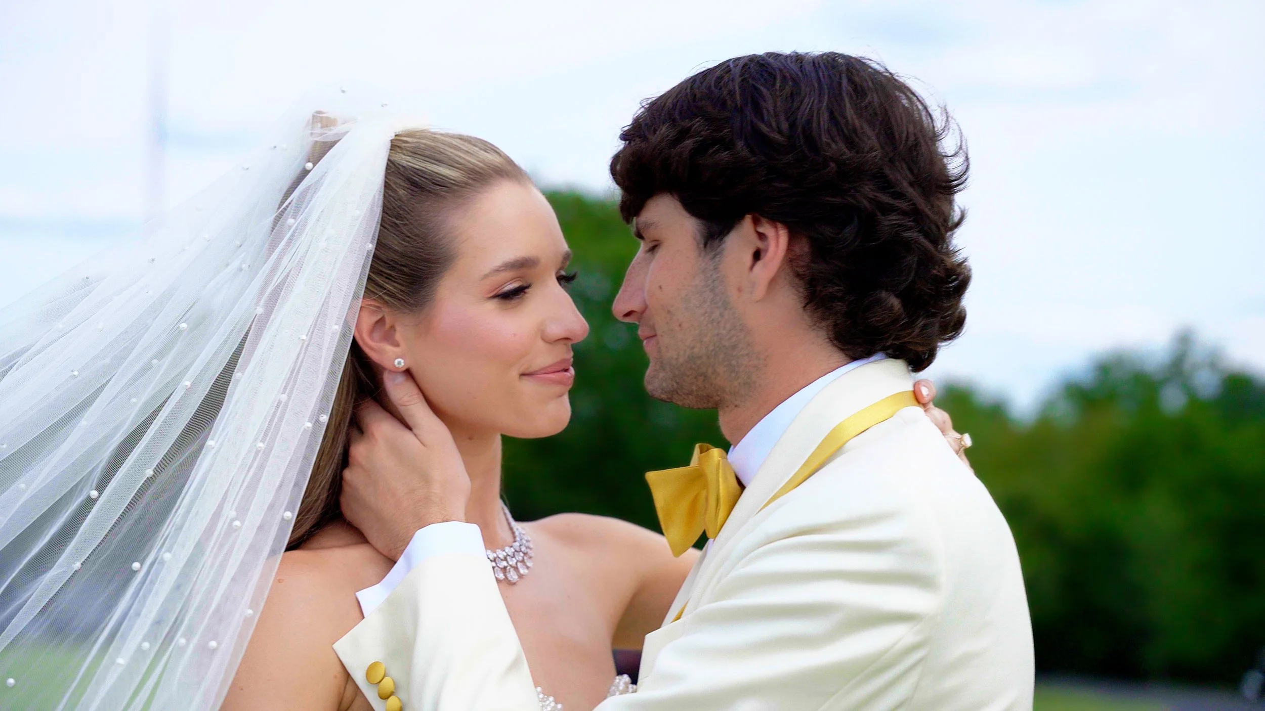 A bride and groom in wedding attire holding each other closely outdoors during daytime, facing each other with smiles.