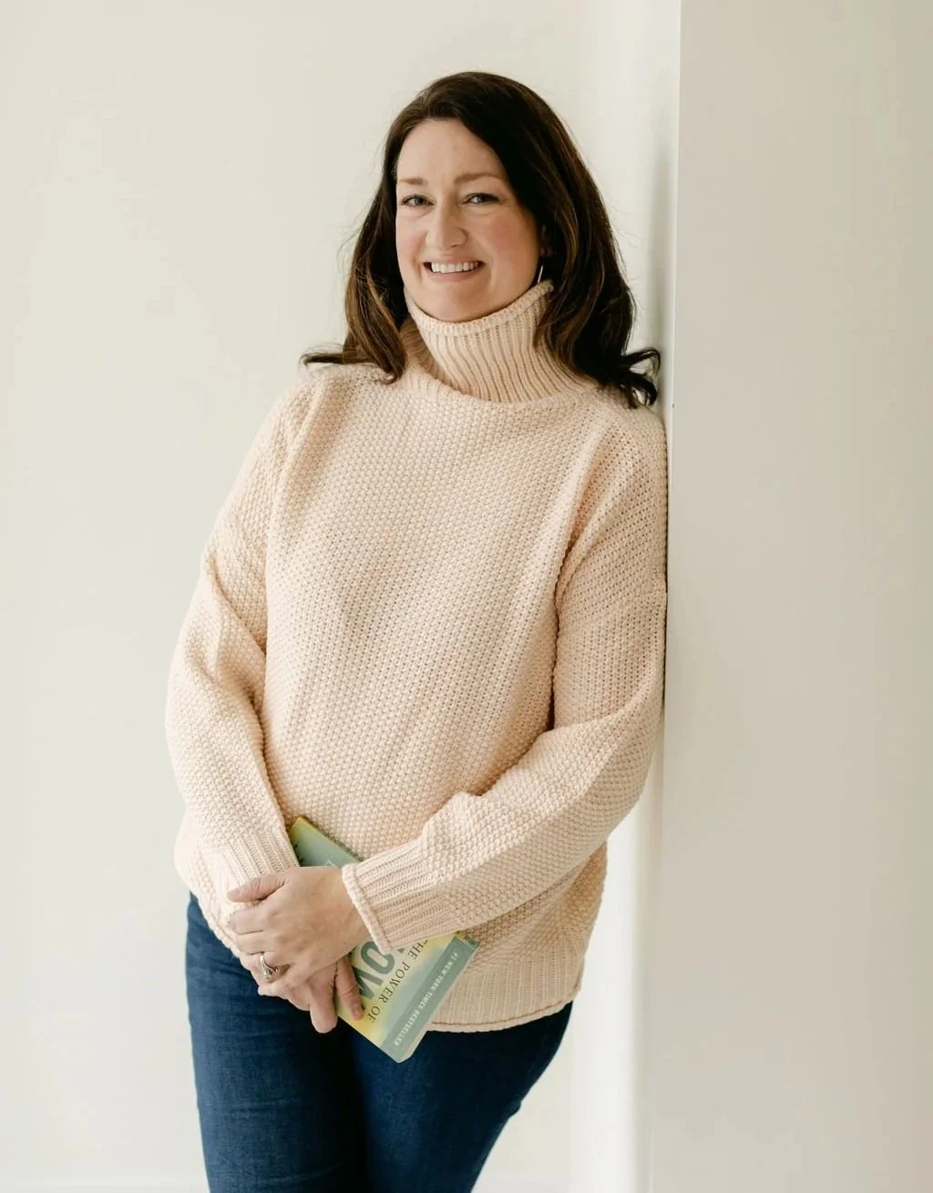 Art Therapist, Emily Davenport and Workplace Wellness Event facilitator in NYC and Westchester. A Workshop led Art therapist woman with dark hair wearing a beige turtleneck sweater and jeans, standing against a white wall and smiling.
