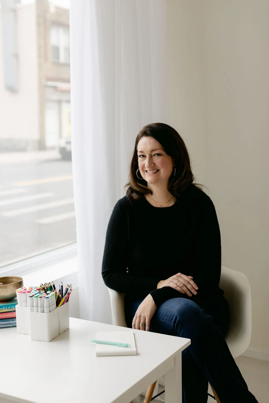 A woman with dark hair smiling and sitting in a bright room with large natural light, white walls, white curtains, a white table, art supplies, and a window with a view of a city street.