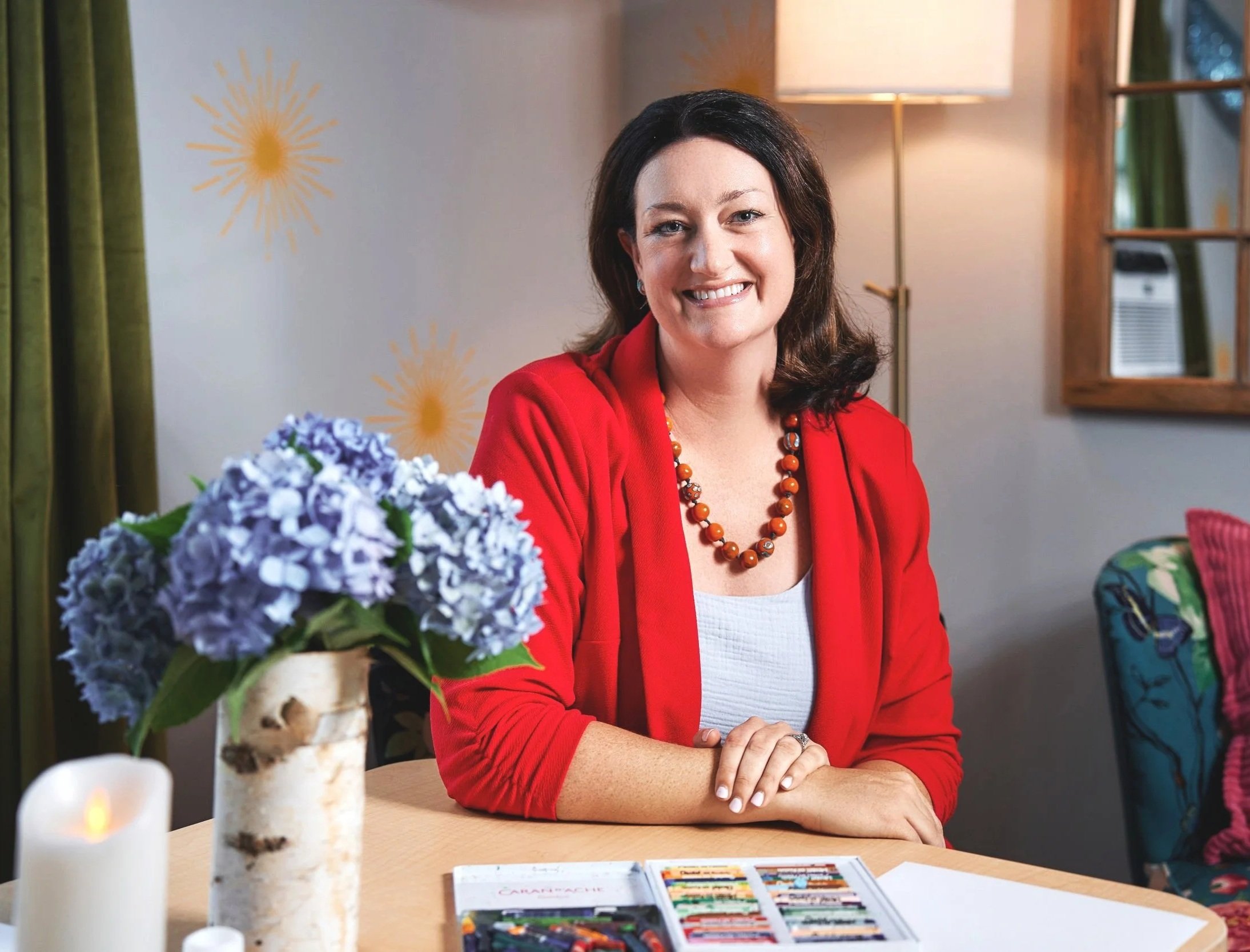 A woman with dark hair, wearing a red blazer and a beaded necklace, smiling with her arms crossed, seated at a table with a bouquet of purple flowers and a candle. The setting appears to be indoors, with a lamp, mirror, and windows in the background.
