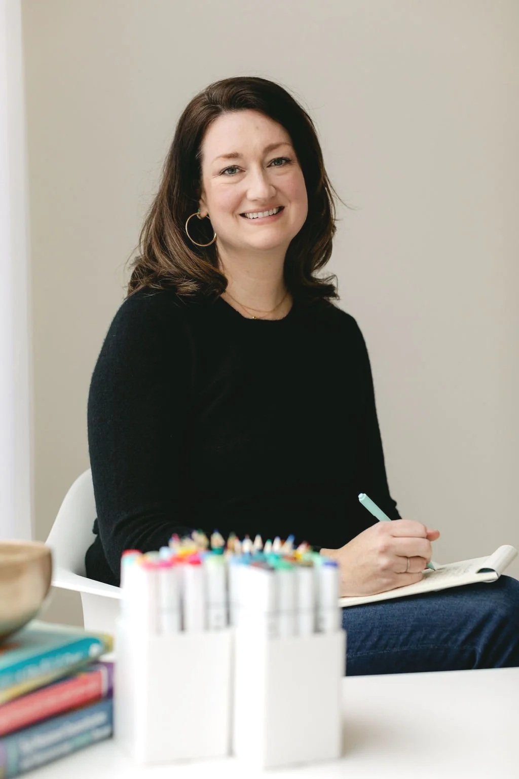 A woman with brown hair, wearing a black sweater and gold hoop earrings, sitting at a table with colored markers and books, smiling at the camera.