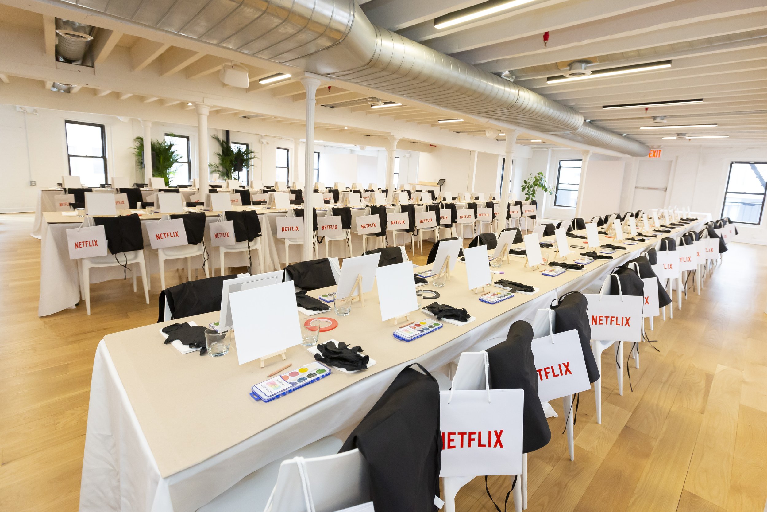 Event setup with tables, chairs, canvas boards, art supplies, and Netflix branded bags in an indoor space.