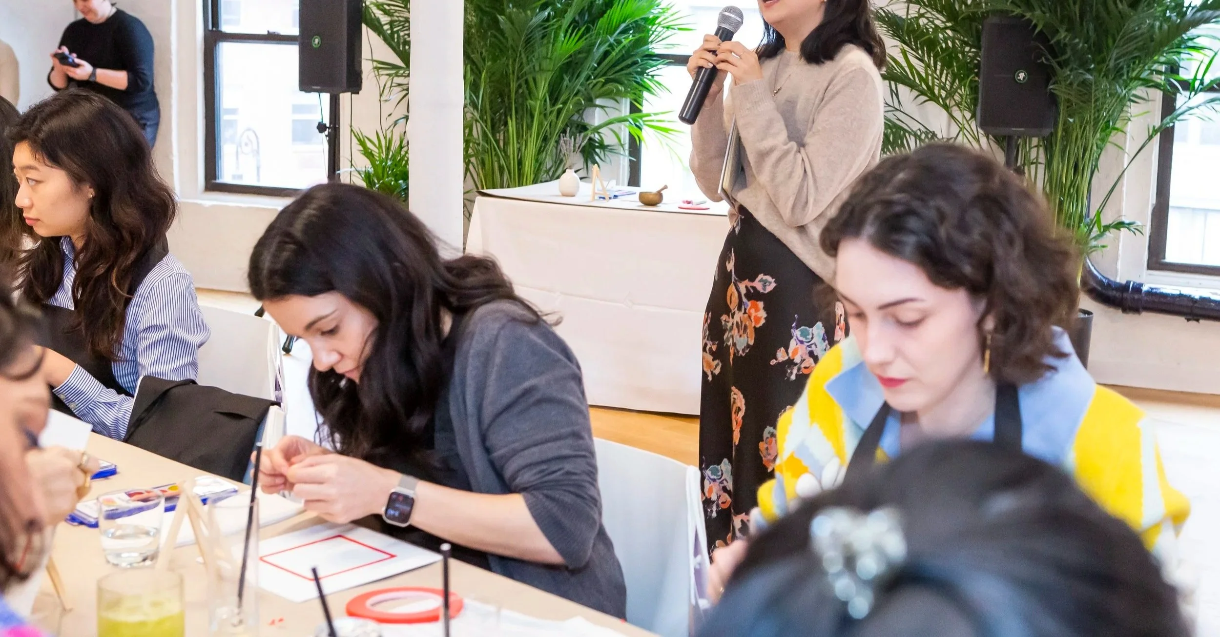 Art Therapy Workplace Wellness Event in NYC. A Workshop led Art therapist standing at a table holding a microphone in a well-lit indoor space, surrounded by several seated women engaged in an activity, with large green plants in the background.