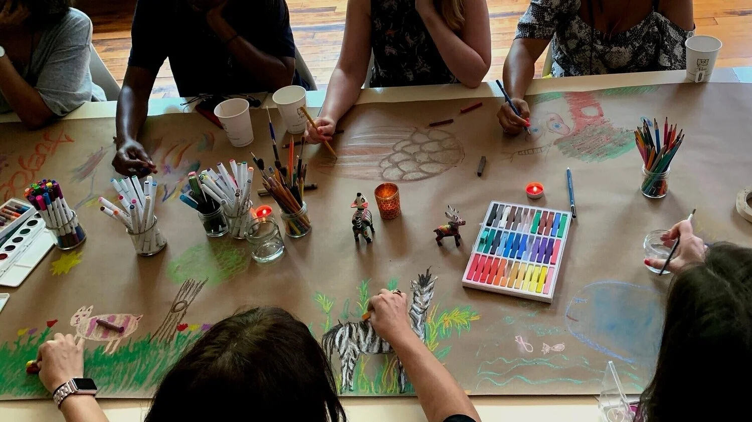 Emily Davenport, Art Therapist, leading teachers in a workshop sitting around a table coloring on a large sheet of paper with various art supplies, including markers, colored pencils, and pastels, with some small decorative figurines and candles. 