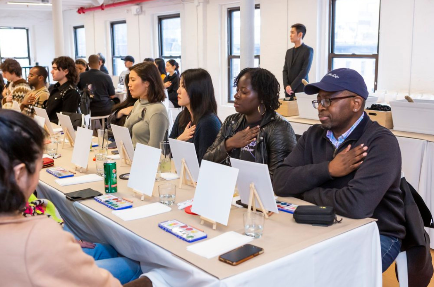 Art Therapy Workplace Wellness Event in NYC. People sitting at a long table with their right hands over their hearts during a pledge or national anthem, in a well-lit room with large windows, with individuals of various ethnicities participating.