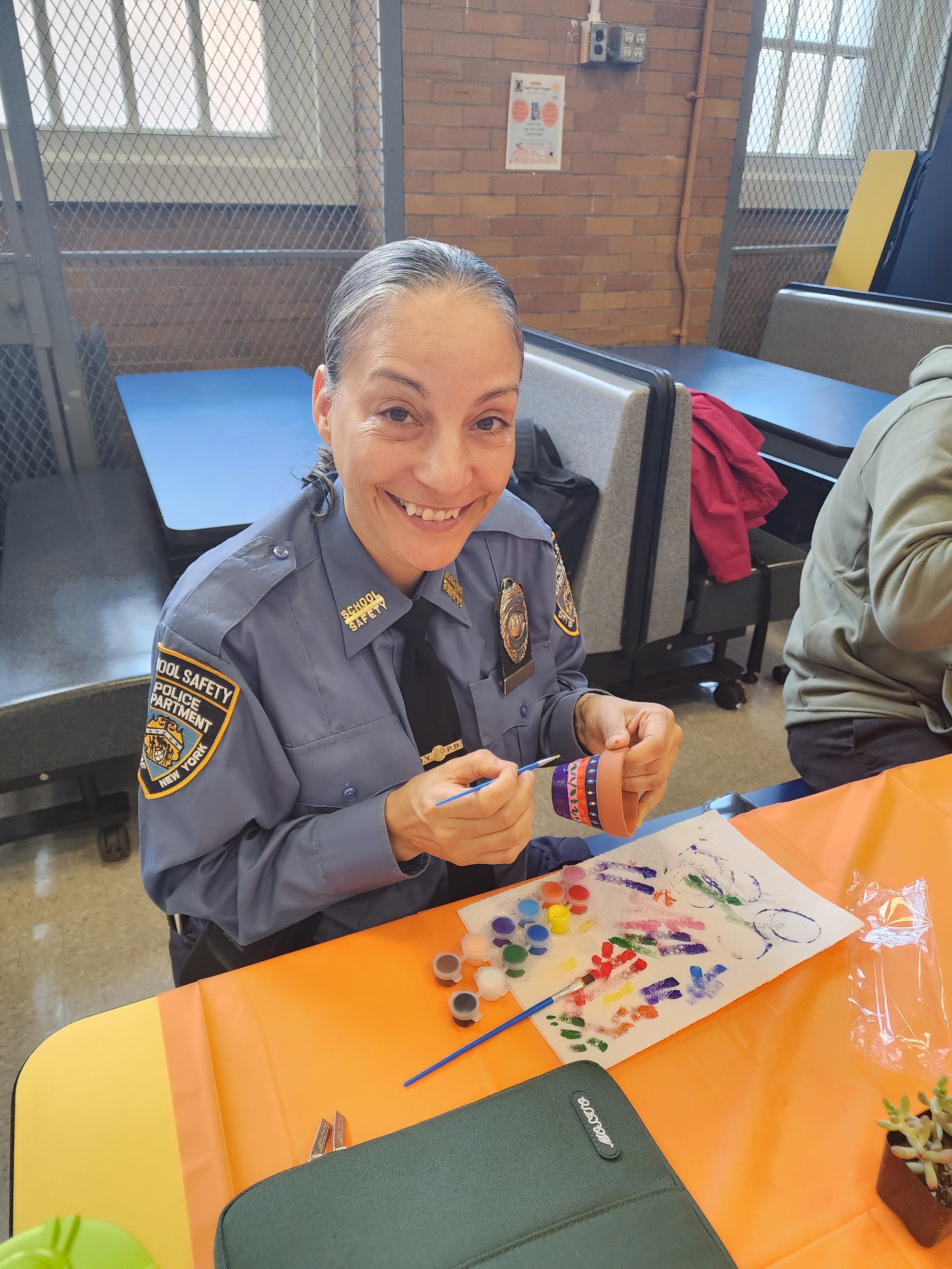 Workplace wellness woman in a police uniform sitting at a table, smiling, painting on a piece of paper with various colors of paint. There are paints, brushes, and a painted paper on the table. The environment appears to be a community center.