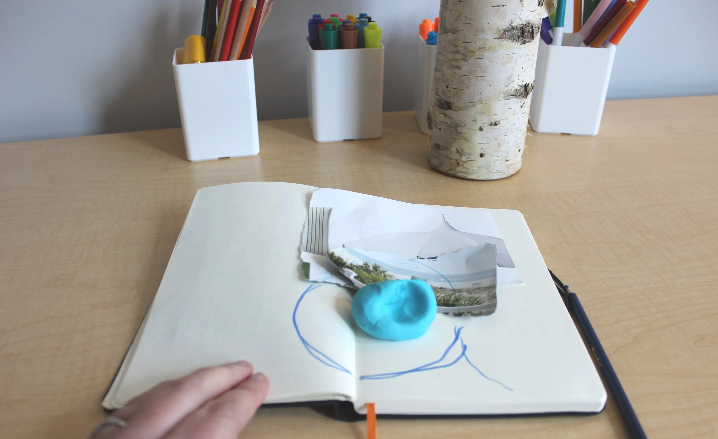 Art Therapy Workplace Wellness Event in NYC. A Workshop led Art therapist modeling an open notebook with pencil sketch of a face, model made of blue modeling clay, and a photograph of a landscape.