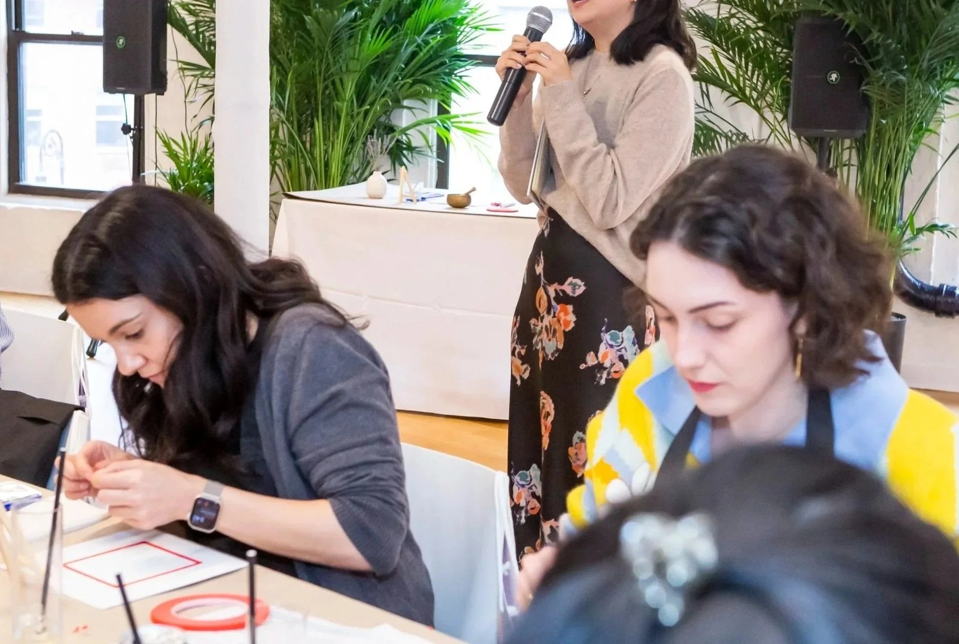 Art Therapy Workplace Wellness Event in NYC led by an Art Therapist in a beige sweater and floral skirt holding a microphone, speaking at an event with two women seated at a table in front of her. The room has plants and black speakers.