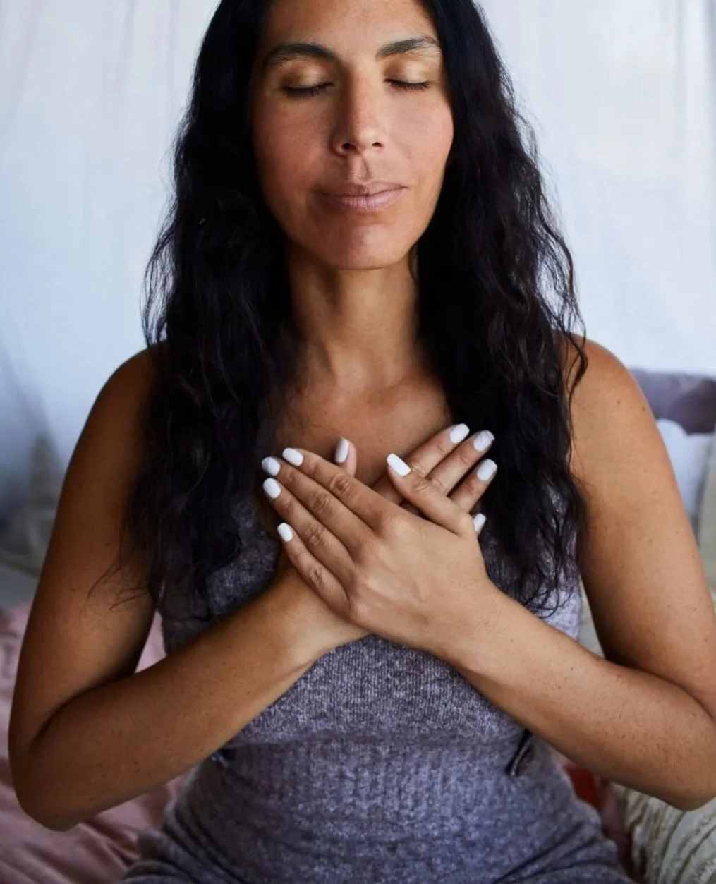 A woman with long dark wavy hair and closed eyes, sitting with her hands on her chest. She has white painted nails and a peaceful expression.