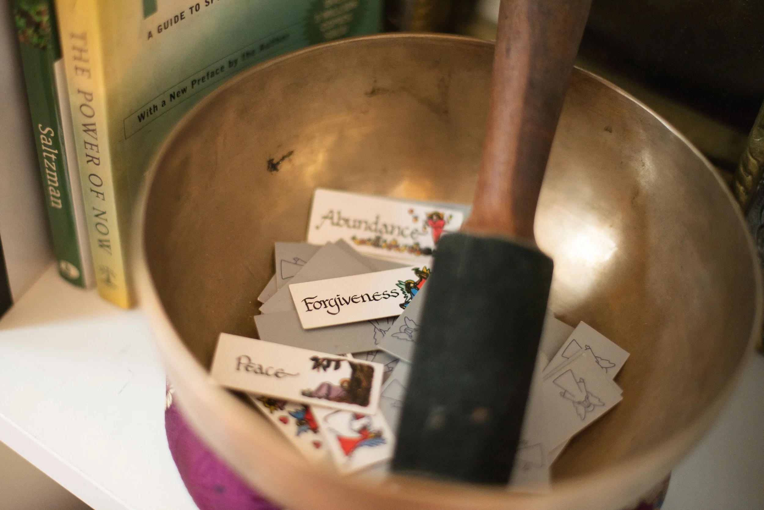 Art Therapist mindfulness singing bowl Inside a metal container with scattered cards, some reading "Forgiveness" and "Peace," and a visible book on the side about a guide to spiritual healing.
