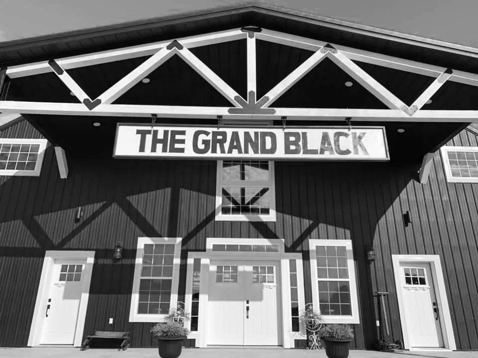 Black-and-white photo of the front entrance to The Grand Black event venue, featuring a large sign with the venue's name above white double doors on a modern black barn-style building.