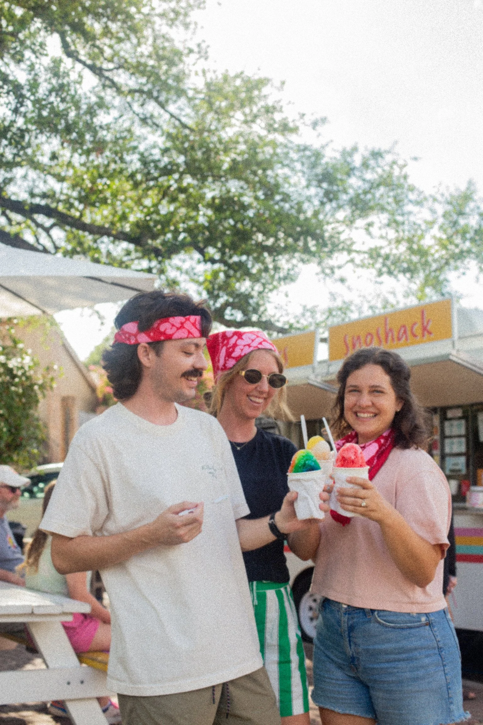 Three friends with pink bandanas enjoying ice cream at an outdoor fair, with a food truck in the background.