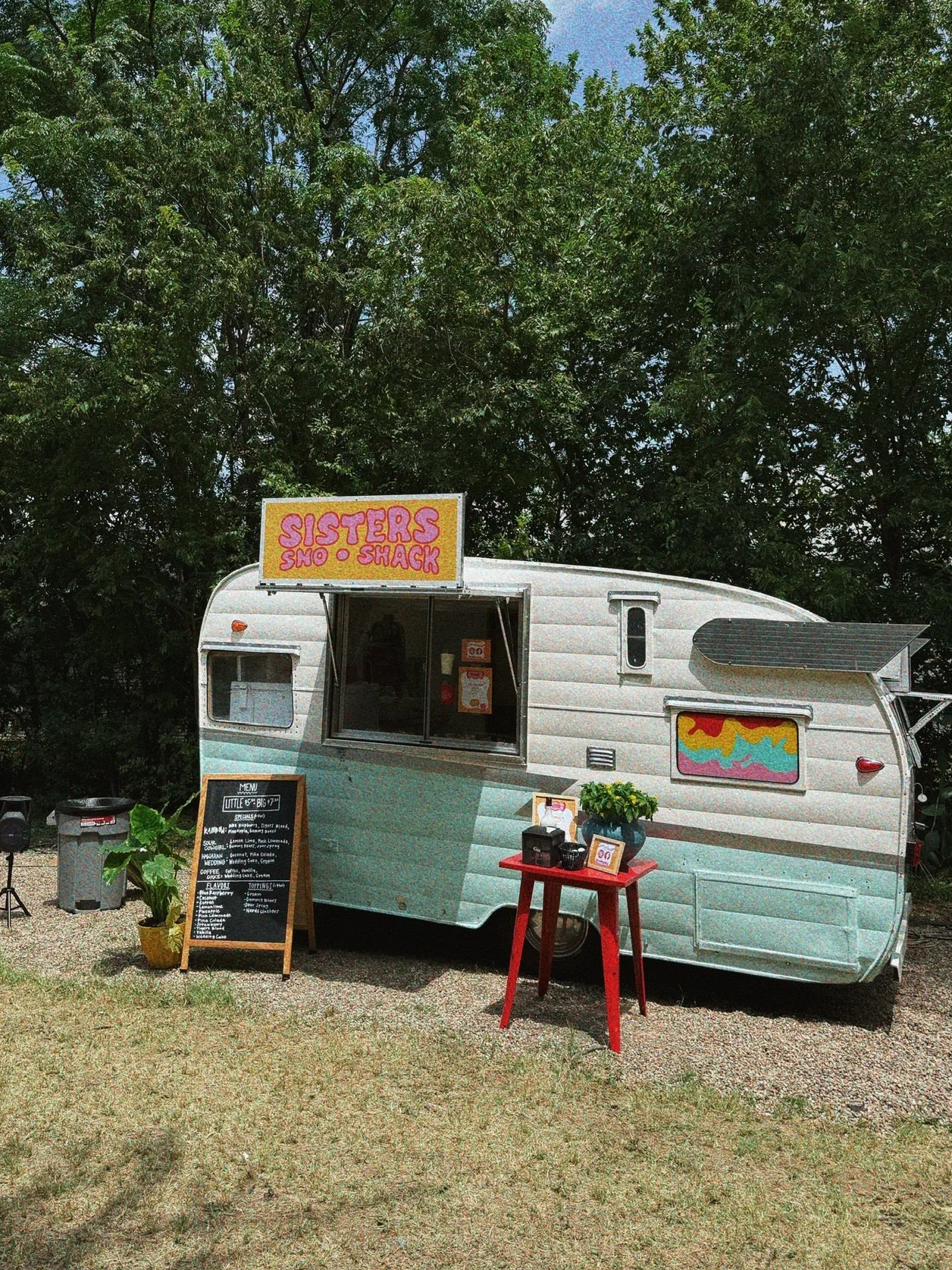 Vintage food truck with a sign that reads 'SISTERS SNACK SHACK', parked outdoors with trees in the background. There is a red table with potted plant and menu items, and a chalkboard menu on the ground.