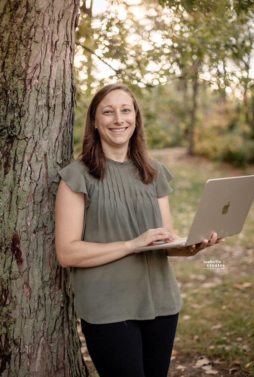 A woman in a green shirt leaning on a tree holding her laptop in park in Noblesville, Indiana.
