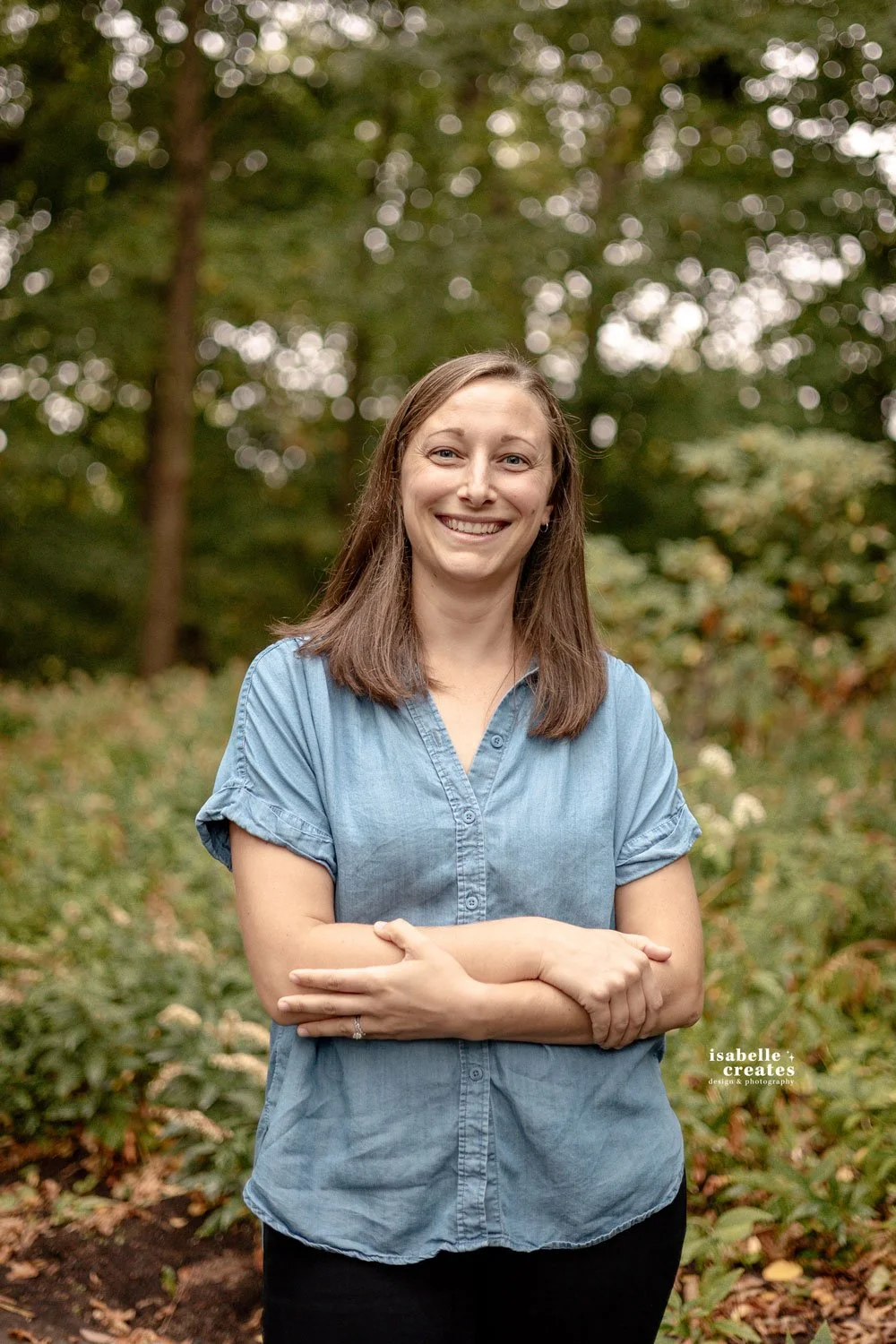 Woman standing with her arms loosely crossed with trees and greenery behind her.