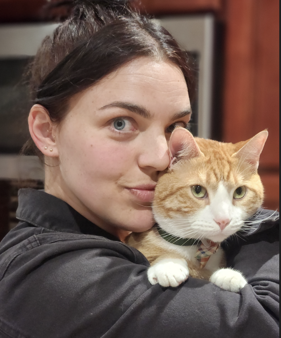 A woman with blue eyes and dark brown hair holds an orange and white cat close to her face.