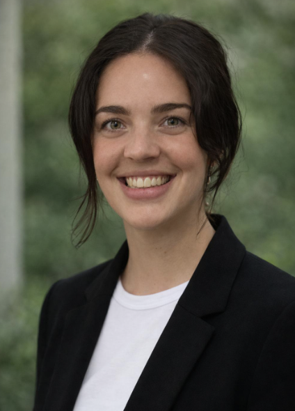 Portrait of a young woman with dark hair in a professional black blazer and white shirt, smiling against a blurred outdoor background.