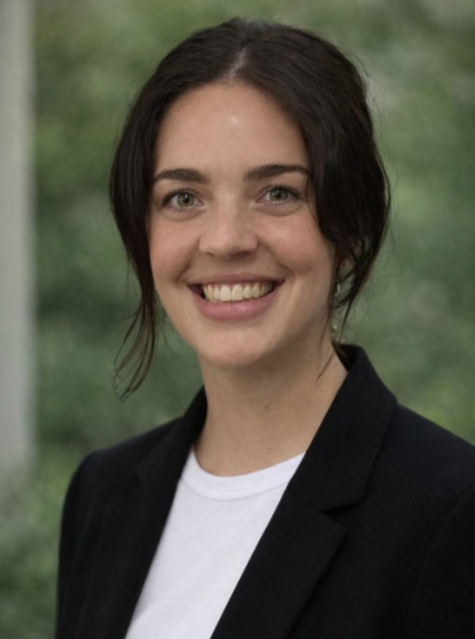 A young woman with short dark hair, wearing a black blazer and white shirt, smiling at the camera outdoors with a blurred green background.