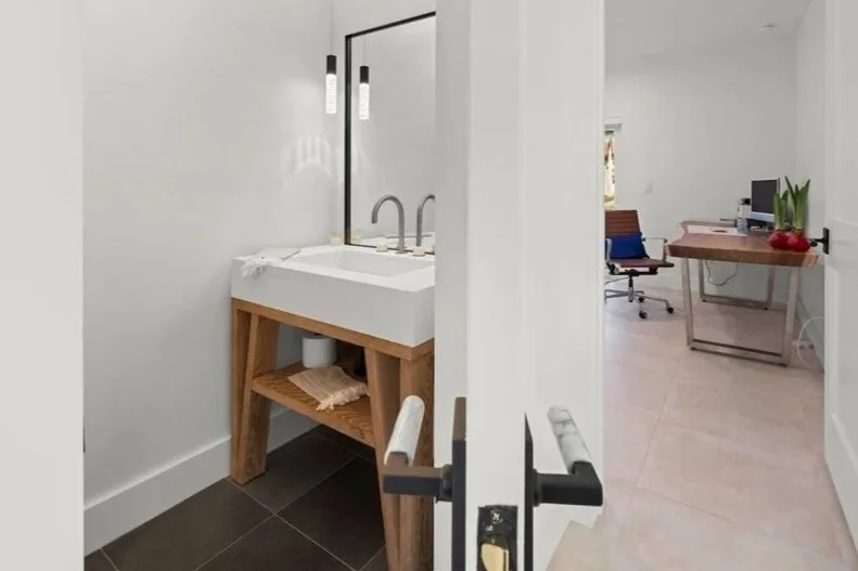A bathroom with a white sink on a wooden stand, and a doorway leading to a home office or workspace with a desk, chair, and computer in a well-lit room.
