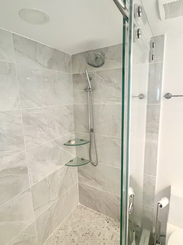 Modern shower with beige and white marble tiles, a chrome showerhead, a handheld shower wand, and two glass shelves.