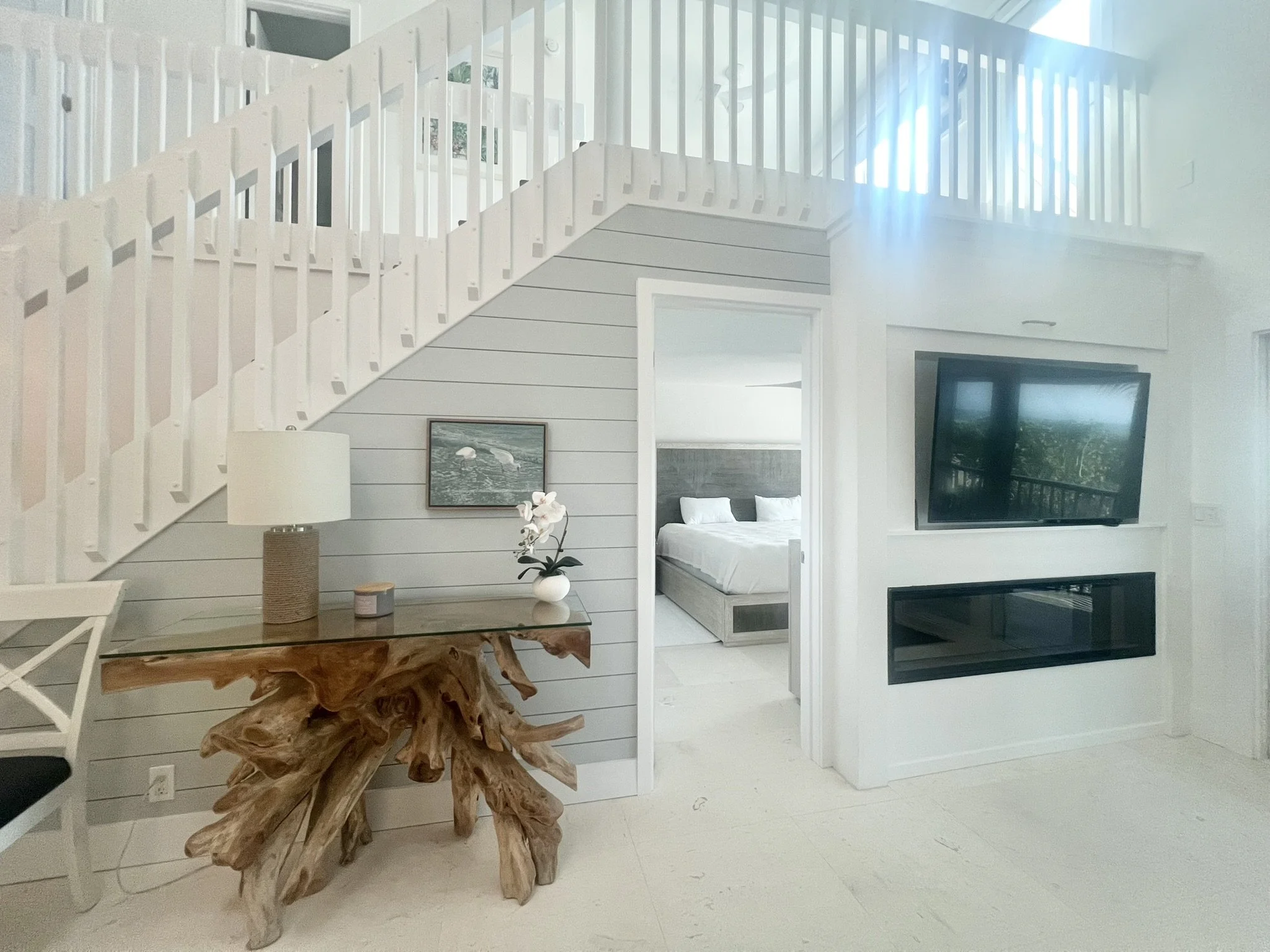 Bright modern living room with a staircase, a wooden table with a lamp and orchid, and a view into a bedroom with a bed and gray headboard.